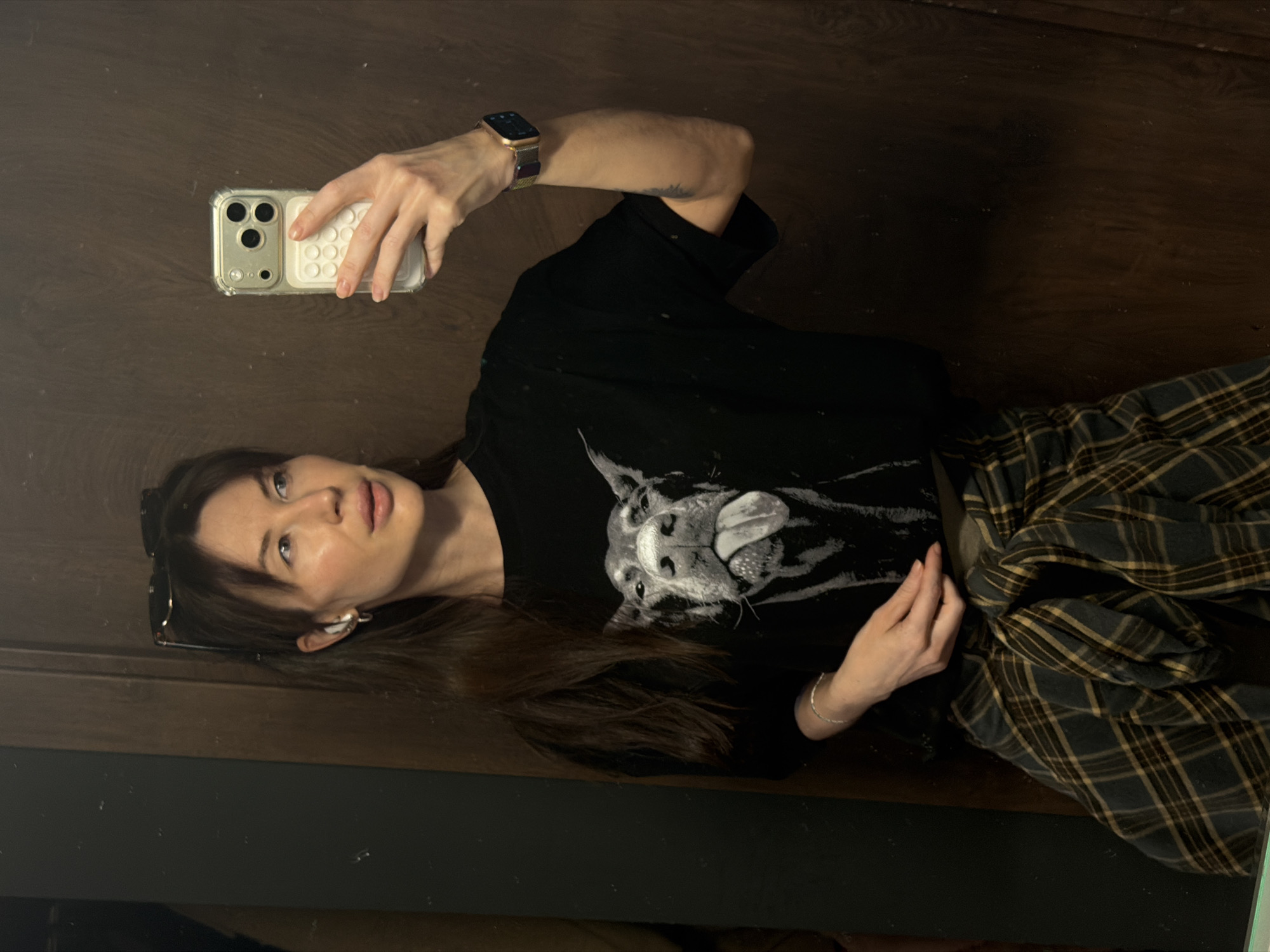 A young woman is seen taking a selfie in what appears to be a mirror. She is wearing a black t-shirt with a dog print, a plaid skirt, and an Apple Watch with a colorful band. She holds an iPhone with a white case in her right hand. Sunglasses are perched on her head. The background consists of what looks like a dark wooden panel. There is no evidence to indicate the specific location in Perm, Russia.