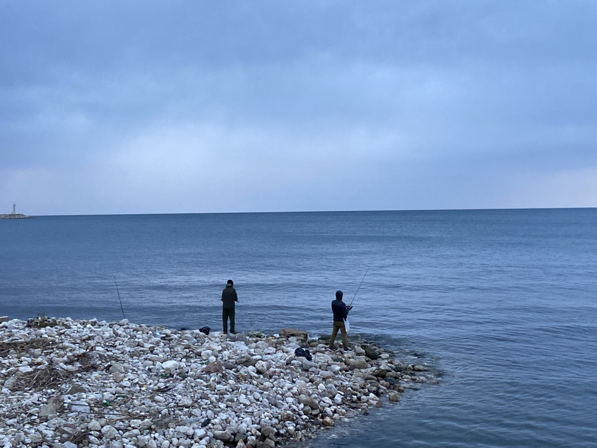 Two individuals are fishing along the rocky coastline in Bar, Montenegro. One person stands in the water, while the other stands on the rocks. Each holds a fishing rod. In the background, across the water, is a small structure that appears to be a lighthouse. The sky is overcast. The shoreline is covered in white stones.