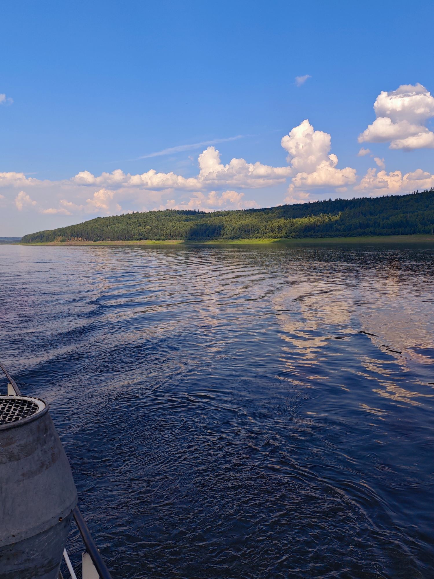 The image depicts a wide, dark blue river flowing towards a lush, green forested hill under a bright blue sky with scattered white clouds. The foreground shows a portion of a boat, including a metal grate and railing. The water has visible ripples and wakes, suggesting movement. The forested hill is dense with trees and rises gently from the riverbank. The sky is clear and bright, with the sun illuminating the clouds and casting a warm light on the scene, particularly reflecting on the water's surface. The location is the city of Podkamennaya Tunguska, Russia, and the overall scene captures a serene natural landscape from the perspective of someone on a boat.