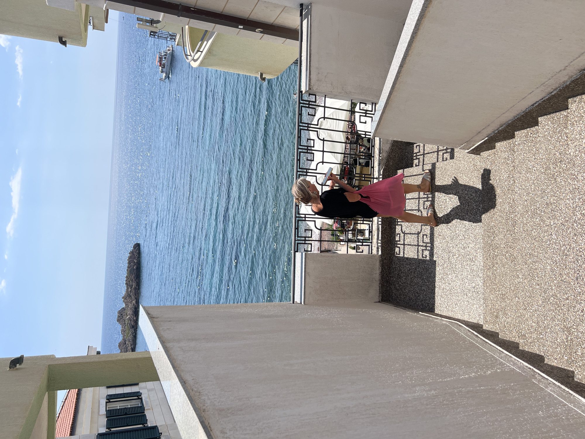 A woman with blonde hair pulled back in a bun, wearing a black dress and flip-flops, walks down a set of outdoor stairs. She carries a bright pink reusable shopping bag in her right hand and holds a phone or small rectangular object in her left hand. She casts a shadow on the speckled concrete stairs. The stairs are bordered by a white wall on the left and a railing with a decorative metal pattern on the right. Beyond the railing, the blue Adriatic Sea is visible under a clear sky with scattered clouds. A small rocky island is situated in the distance, and a boat with people on it is moving across the water. The scene appears to be at a seaside resort or hotel in or near Sveti Stefan, Montenegro, with a balcony and part of a building visible on the right side of the frame.