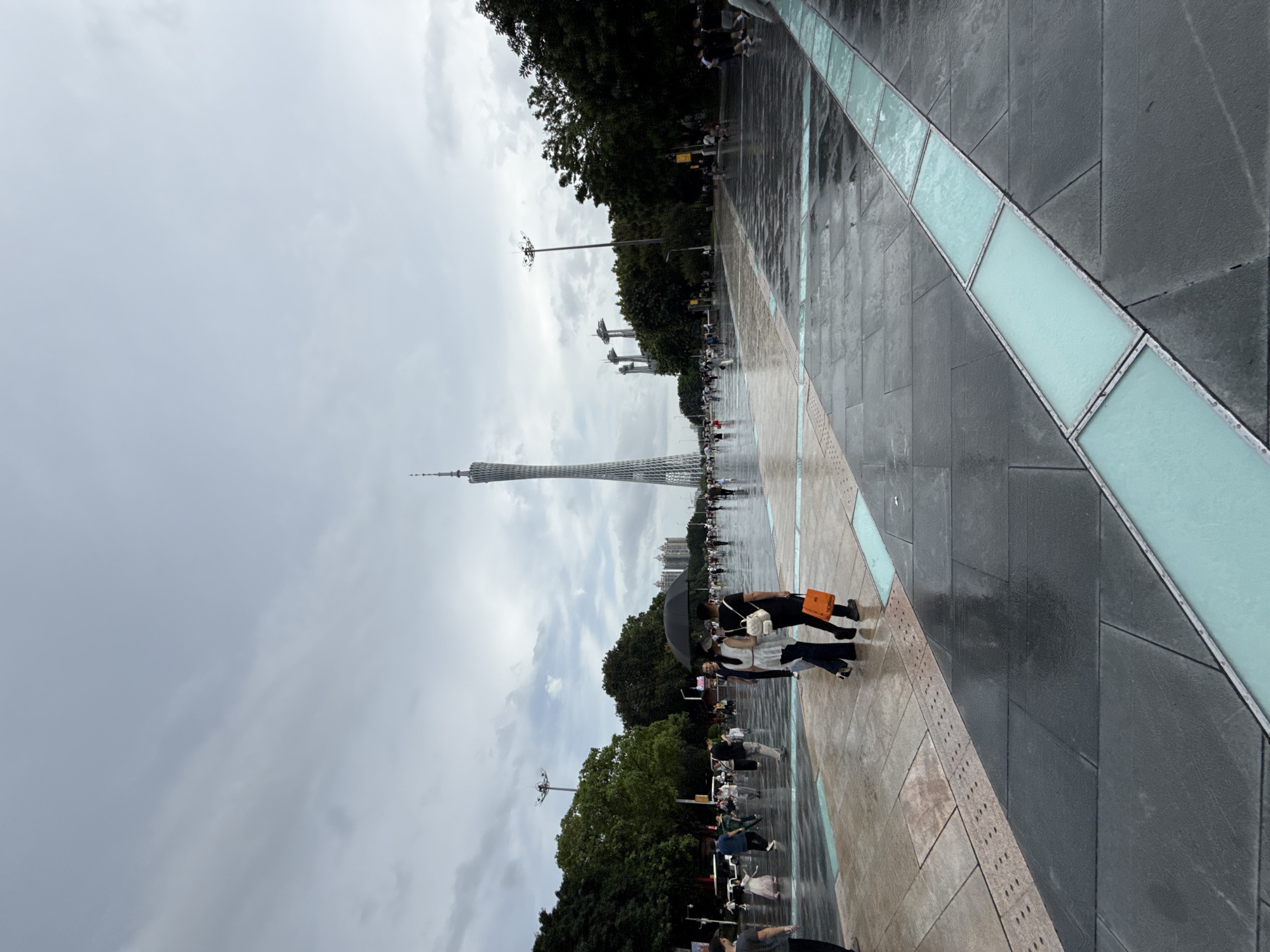 The image depicts an outdoor scene in Tianhe District, China, with the Canton Tower prominently visible in the distance. The ground is wet, suggesting recent rain, and people are walking on a paved area. Several individuals are walking together, with one person holding a black umbrella. In the foreground, there is a patterned pavement with sections of light blue, translucent material. Trees line the sides of the area, and streetlights are also present. The sky is overcast with gray clouds.