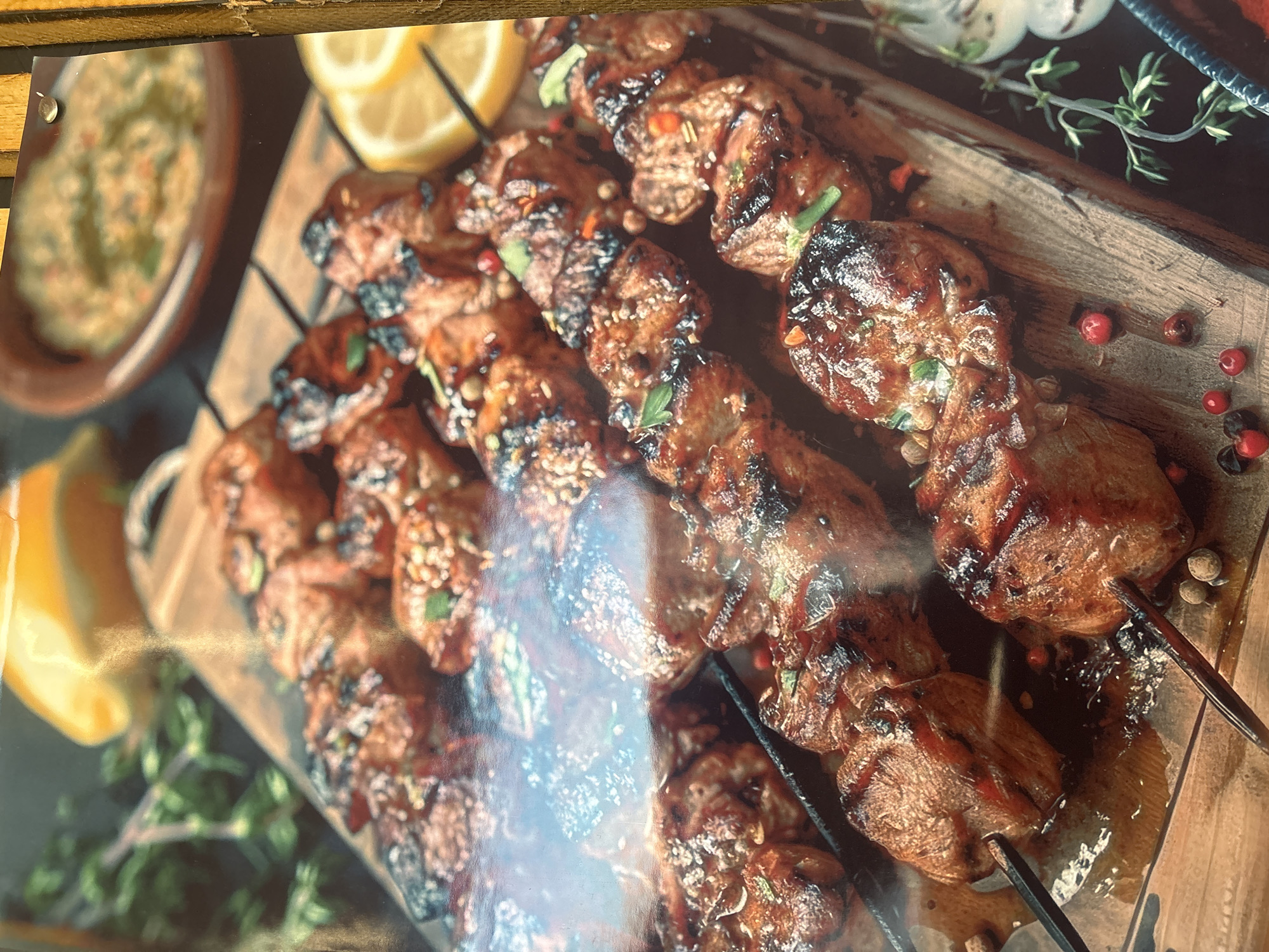 The image depicts a platter of grilled meat skewers served on a wooden board. The meat appears to be glazed and garnished with herbs and spices. Lemon wedges and a bowl of sauce are also present. A sprig of thyme and scattered peppercorns decorate the board. The location is unknown, and there's no information suggesting it's Krasnoyarsk, Russia. The image doesn't include people or their interactions.