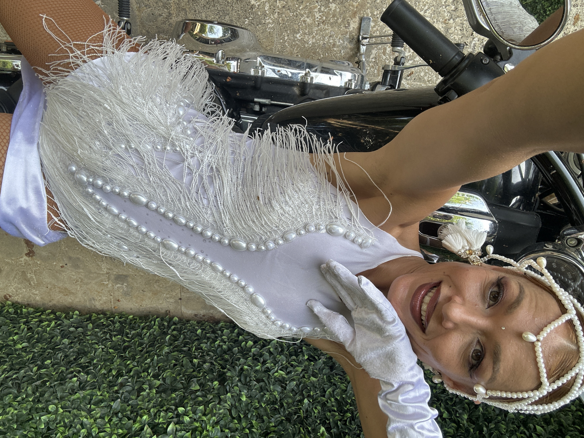 A woman in a white, fringed costume and pearl headpiece, complete with white gloves, leans back on a black motorcycle. She is positioned outdoors with a green hedge and a concrete ground as the backdrop. The scene appears to be set in a public space, possibly a street or a pathway, in the city of Ko Samui District, Thailand. The woman's attire suggests a festive or performance-oriented occasion.