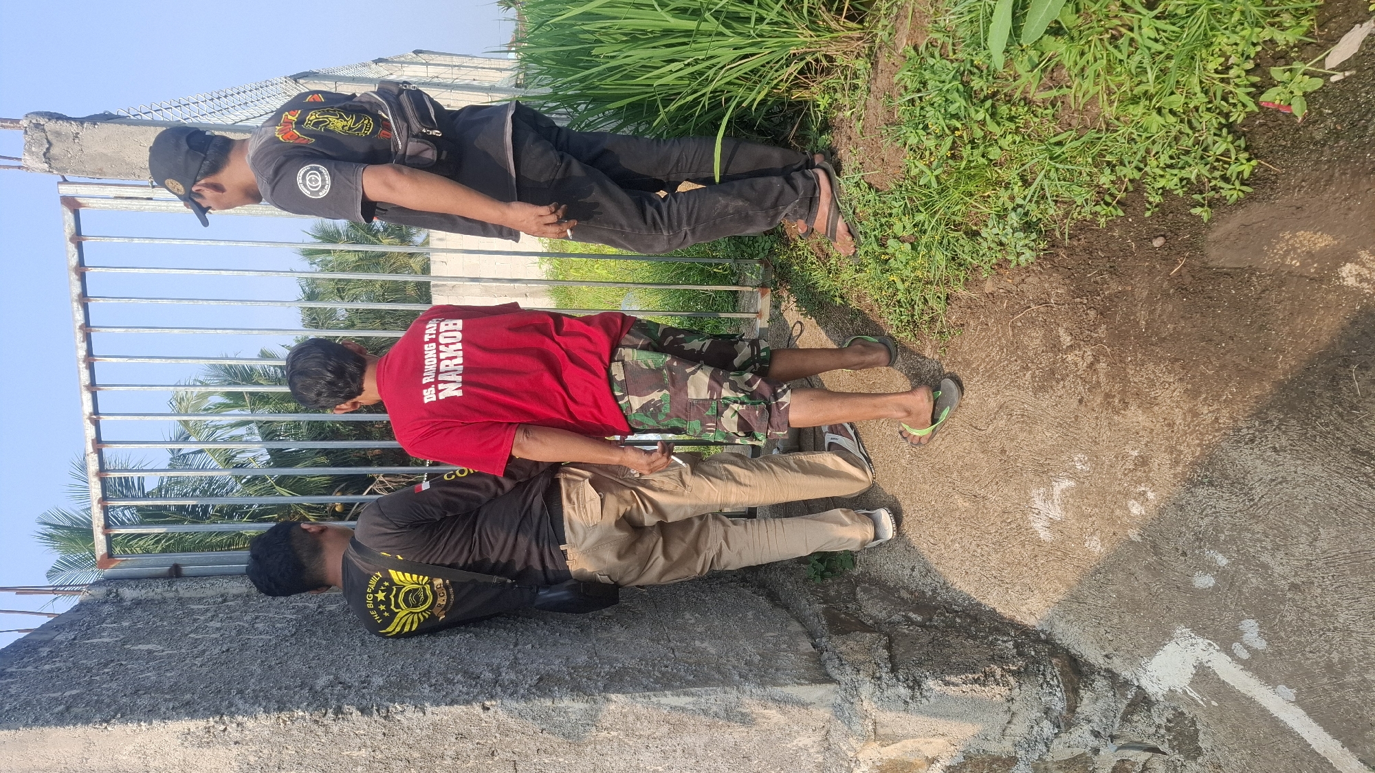 Three males are standing near a metal gate, with a concrete wall to their left and a field of green vegetation to their right. The man on the far left, wearing a black t-shirt with a yellow emblem and a black crossbody bag, is leaning against the concrete wall, facing away from the camera. He is wearing khaki-colored cargo pants and white sandals. The man in the center, wearing a red t-shirt with white text that reads "DS. RAHONG TANAH NARKOBA," is facing away from the camera, looking towards the gate. He is wearing camouflage cargo shorts and green flip-flops. He is holding a cigarette in his right hand. The man on the far right, wearing a black t-shirt with a circular emblem on the back, a black baseball cap, and black jeans, is also facing away from the camera, looking towards the gate. He has a small black bag slung across his chest and is wearing black flip-flops. He is also holding a cigarette. The environment appears to be outdoors, with a clear blue sky visible. The ground is a mix of concrete and dirt, with grass and other vegetation growing to the right of the gate. The scene suggests a casual gathering or a moment of pause in an outdoor location. The text on the red shirt indicates the location is in or related to "Rahong Tanah Narkoba," which could be a village or area in Indonesia.