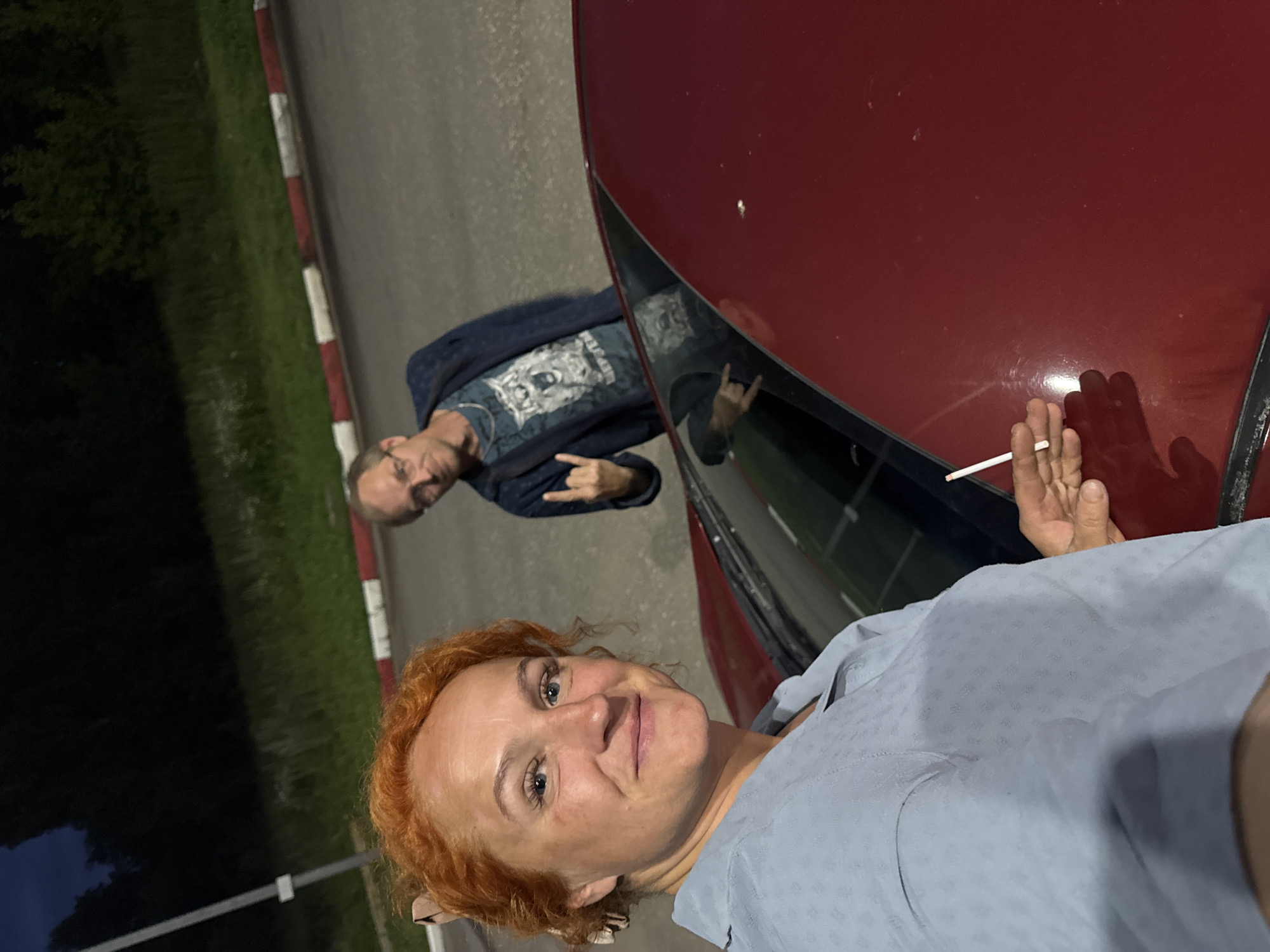 The image features two people near a red car at night. The person in the foreground is a woman with short, reddish-blonde curly hair and blue eyes, smiling at the camera. She is wearing a light blue collared shirt with a subtle textured pattern. Her right hand is visible, holding a lit cigarette. The background shows a man with short hair and glasses, wearing a dark blue denim jacket over a t-shirt with a graphic print. He is looking towards the camera and has his left hand raised, forming a "rock on" gesture with his fingers. The scene appears to be outdoors, possibly a roadside or parking area, with a dark grassy area and a road visible. A curb with red and white markings is also present. The lighting suggests it is nighttime. The overall mood seems casual and relaxed.