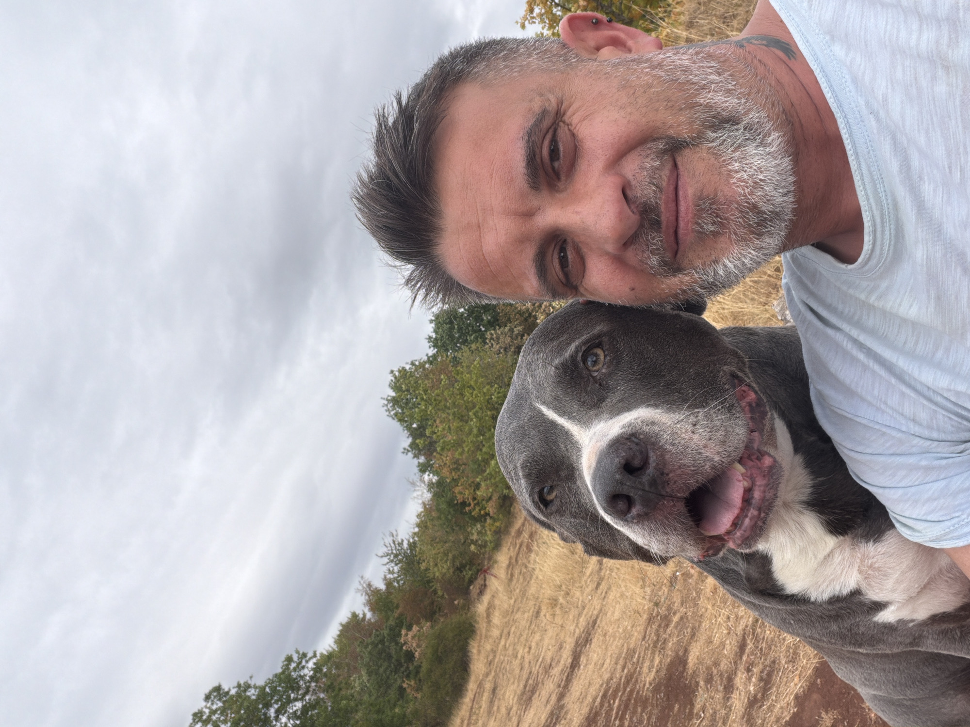 A man and a dog are posing for a selfie outdoors. The man, with short, spiked gray hair and a salt-and-pepper beard, is smiling and looking at the camera. He has a tattoo on his neck and an earring in his left ear. To his left is a large gray dog with white markings on its chest and face, panting with its tongue out. The dog's head is turned towards the man. They are positioned on a dry, grassy hillside with trees in the background under a cloudy sky. The location is not specified as Esch-sur-Alzette, Luxembourg.