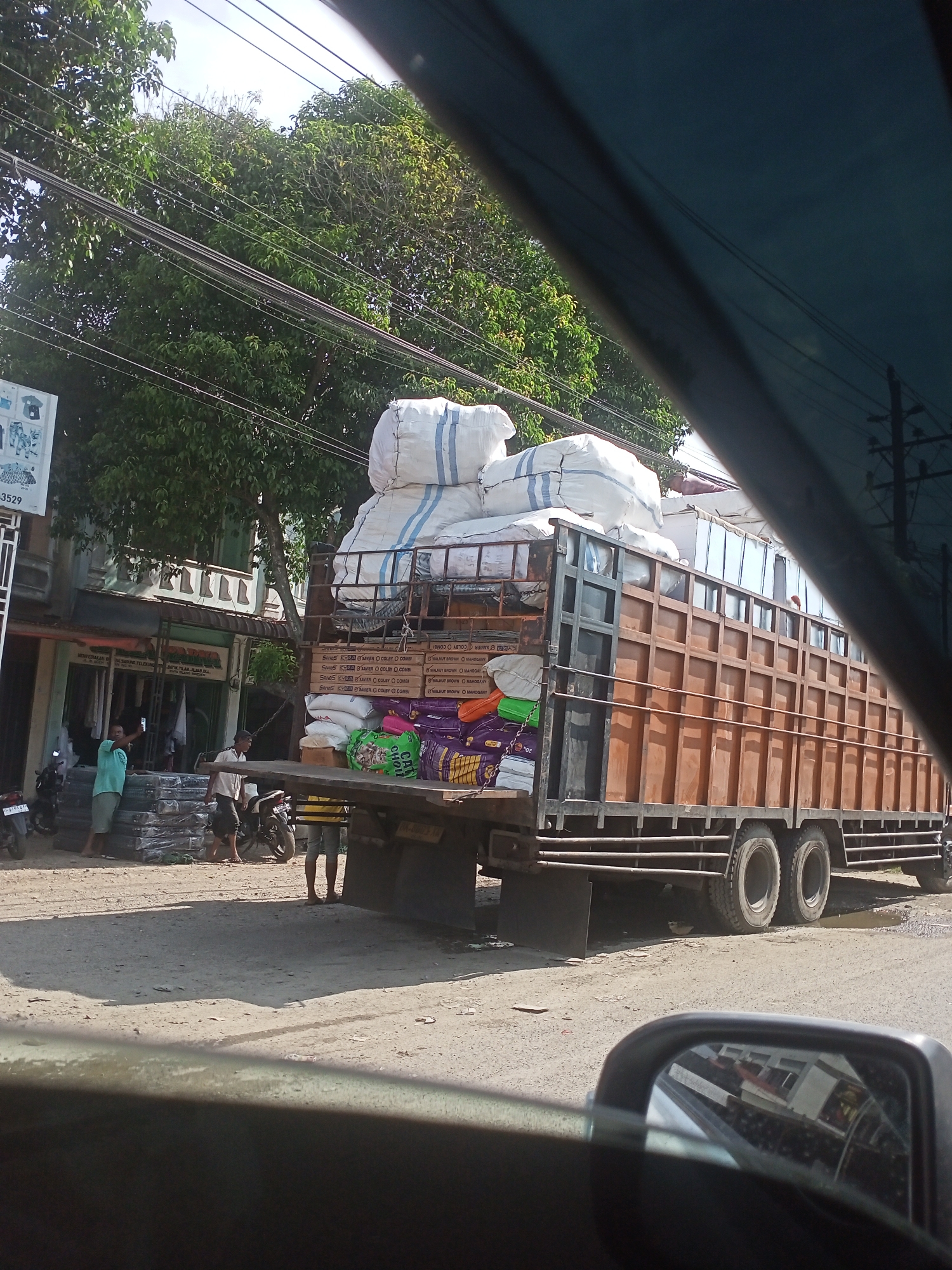 This image captures a bright daytime street scene in Tingkeummanyang, Indonesia, from the perspective of a vehicle's passenger seat. The foreground shows part of a car's dashboard and a side mirror reflecting some buildings.
The main subject is a large brown truck, heavily laden with various goods. On the top, several oversized white sacks are stacked, secured to the truck's frame. Below these, multiple cardboard boxes are visible, displaying text such as "XAVIER COLBY COMBI" and "WALNUT BROWN MAHOGANY", suggesting furniture or building materials. Further down, colorful bags, including a prominent green one with "CAT FOOD" partially legible, and a purple one, are also part of the cargo.
Near the rear of the truck, several men are present on a dirt or unpaved road. One man in a light blue shirt stands by a stack of black crates or panels, possibly supervising or participating in loading/unloading. Another man, wearing a white shirt and dark shorts, is also near the goods. A third man is partially visible further left. Two motorcycles are parked nearby.
In the background, a building with a green awning and intricate architectural details is visible. Lush green trees and numerous power lines crisscross the sky, indicating a typical semi-urban or rural setting. The weather is sunny with clear skies, casting strong shadows on the ground.
Visible text includes "-3529" on a sign on the building to the left, and various product names on the cardboard boxes on the truck: "SannS KKB XAVIER COLBY O COMBI", "SS AERO COLBY COMBI", "WALNUT BROWN MAHOGANY", and "WALNUT BROWN MAHOG/Y". A green bag clearly shows "CAT FOOD".