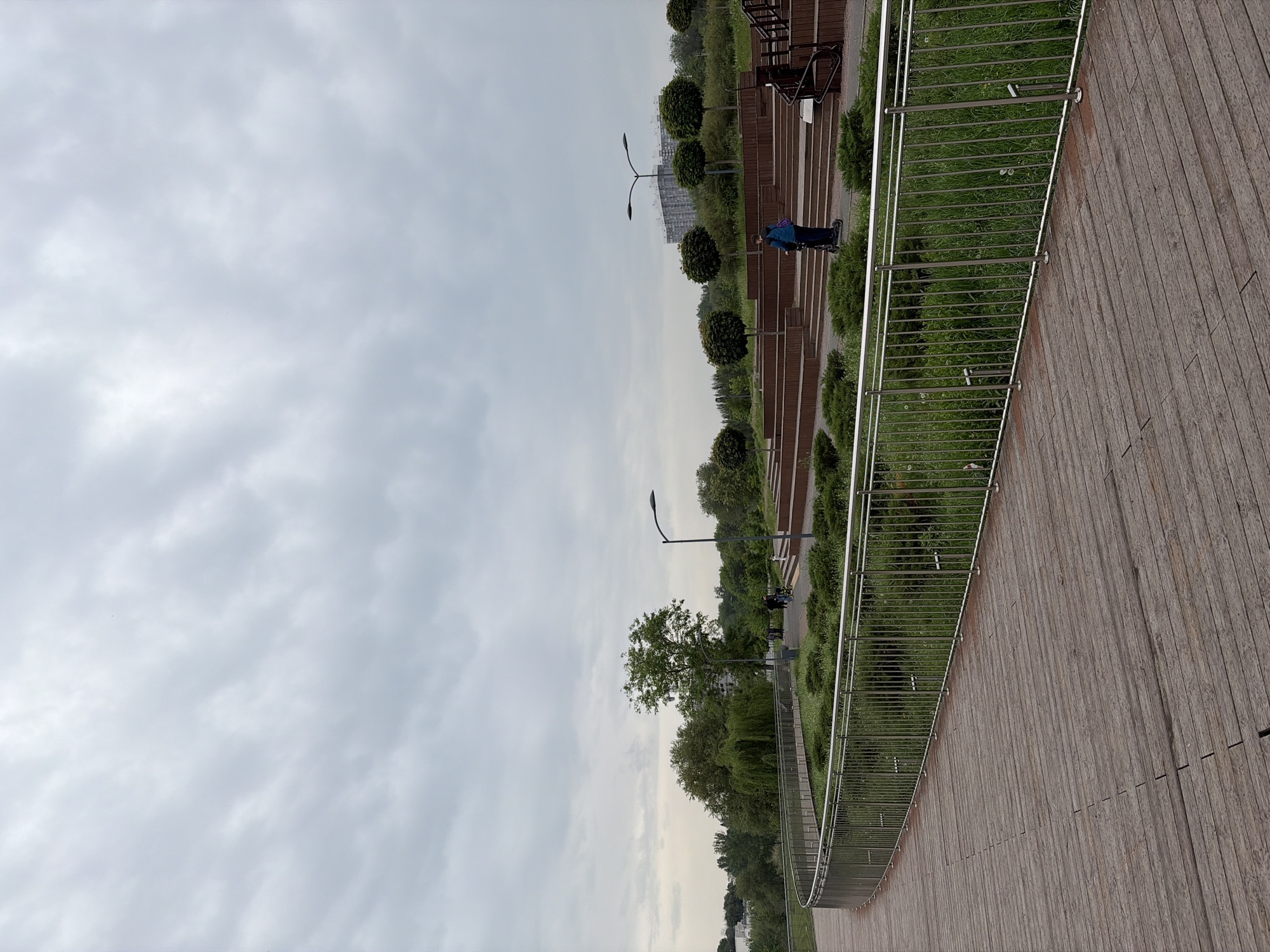 The image captures a view of a park in Rostov-on-Don, Russia, under an overcast sky. A wooden boardwalk with a metal railing occupies a significant portion of the foreground, curving along the edge of the park. Parallel to the boardwalk, a landscaped area features manicured, round bushes and trees. A few benches are visible among the greenery. In the distance, a person wearing a blue jacket stands on a pathway within the park. Street lamps line the path. In the background, there is a glimpse of city buildings and trees, partially obscured by the cloudy sky.