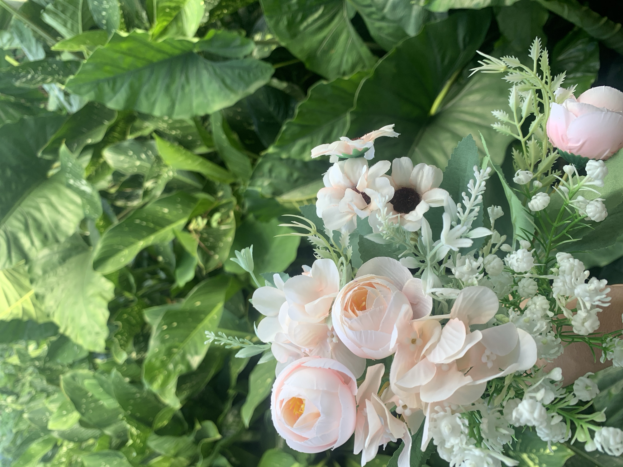 A bouquet of artificial flowers, predominantly in shades of pale pink and white, is held in front of a lush, green leafy background. The bouquet features several rose-like blooms, smaller daisy-like flowers with dark centers, and delicate white filler flowers. The leaves in the background are large and appear to be from tropical plants, creating a dense, verdant setting. The lighting suggests it is daytime. There is no visible indication of the specific location within Kuala Pembuang, Indonesia, other than the general botanical environment.