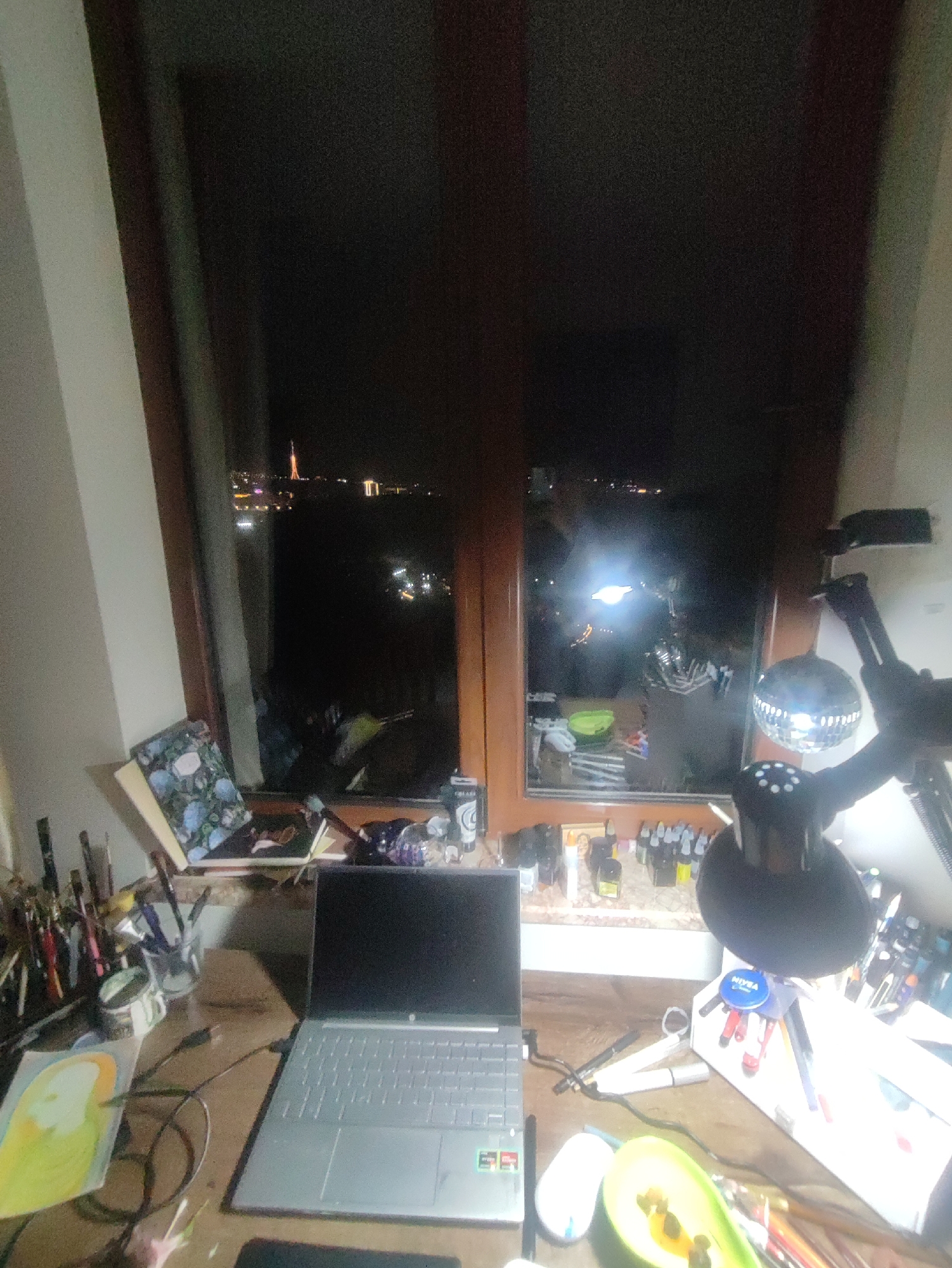 The image shows a desk in front of a window at night. On the desk, there is a laptop, an open avocado, a mouse, paintbrushes, paint, and other art supplies. Outside the window, the city lights of Yerevan, Armenia are visible, including a tall tower-like structure on the left. A disco ball and desk lamp are to the right of the window.