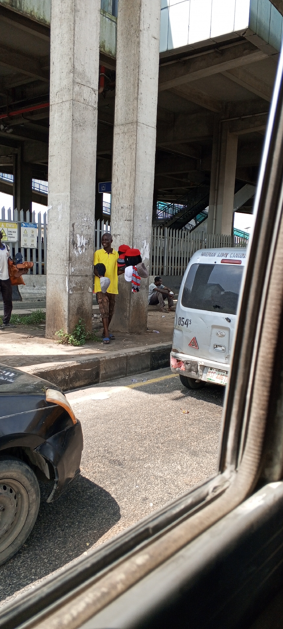 A man in a yellow t-shirt and patterned shorts stands near a large concrete pillar, holding a collection of red and black hats, along with a striped scarf. He appears to be a vendor, possibly selling these items. In the background, a silver van with the lettering "NIGERIAN ARMY CANTEEN" is visible, along with another man sitting on the ground. The scene is outdoors, under an elevated structure with more concrete pillars and railings. A black car is partially visible in the foreground, viewed from inside another vehicle, suggesting the photo was taken from a car window. The ground is asphalt, and there are some weeds growing near the base of the pillar. The environment appears to be a busy urban area in Agani, Nigeria, likely near a transportation hub or market.