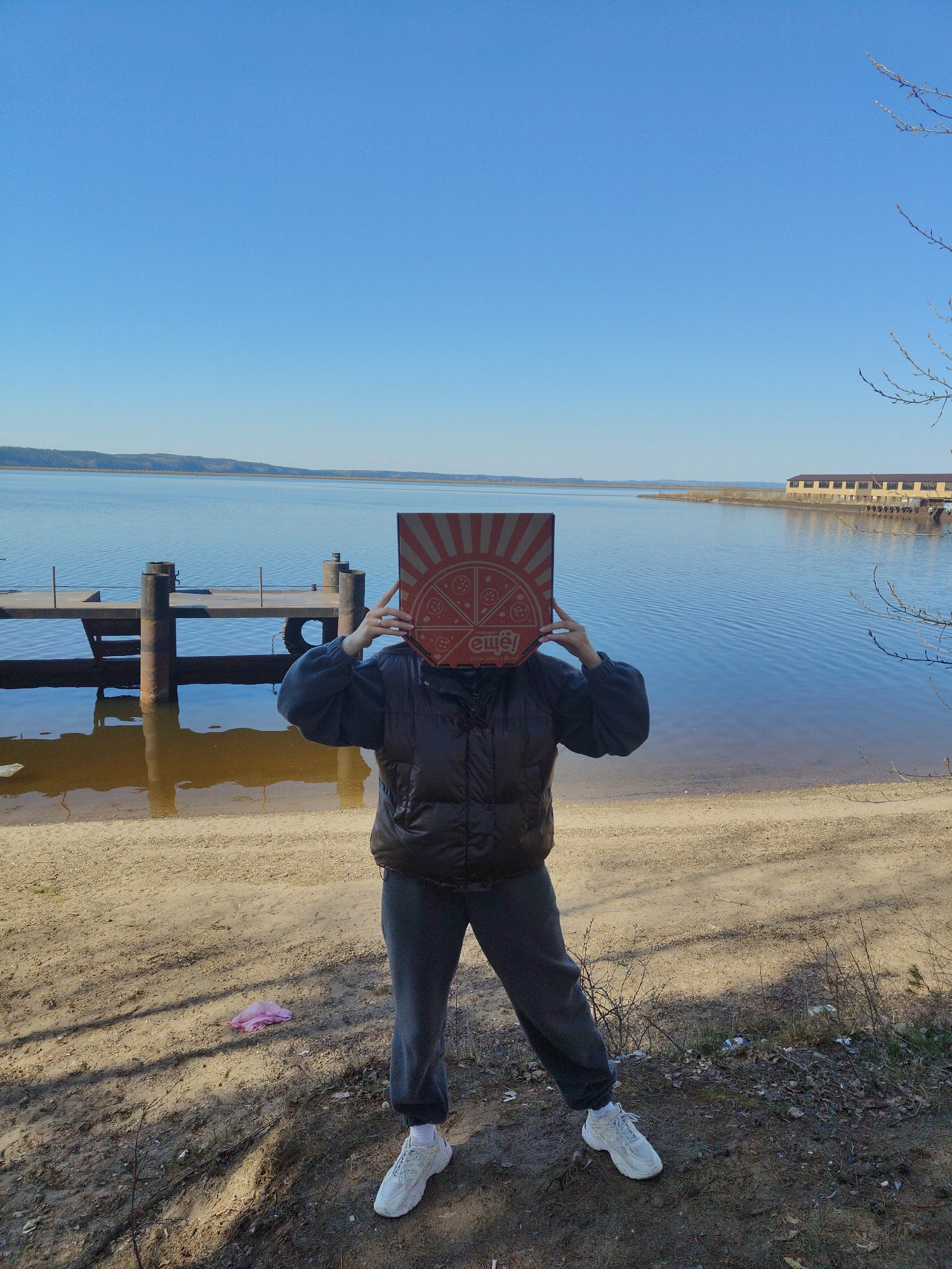 A person stands on a sandy shore next to a body of water, holding a square pizza box in front of their face. The person is wearing a dark puffy vest over a dark sweatshirt, dark sweatpants, and white sneakers. The water stretches to the horizon under a clear blue sky. To the left is a wooden pier, and to the right, a building is visible in the distance. A small, crumpled pink object is on the sand to the left of the person's feet. Sparse vegetation grows on the shore. The location is in the city of Unknown, Unknown.