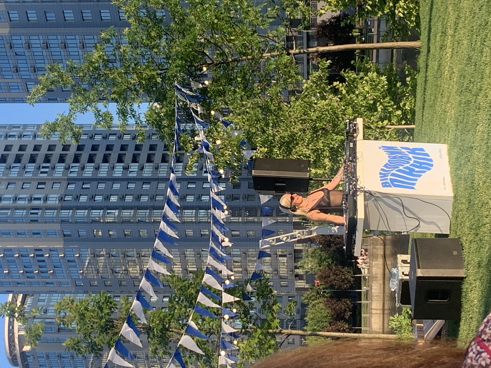 A blonde woman wearing headphones and sunglasses is playing a DJ set outdoors, on a grassy area with a river and modern buildings in the background. She is standing behind a white podium with DJ equipment, and a large speaker is positioned to her left. Decorative blue and white flags are strung across the scene above the DJ. The location is the Obolonska Embankment in Kyiv, Ukraine, specifically near the "River Mall" shopping center. A portion of another person's head and shoulder are visible in the foreground on the left side of the image.