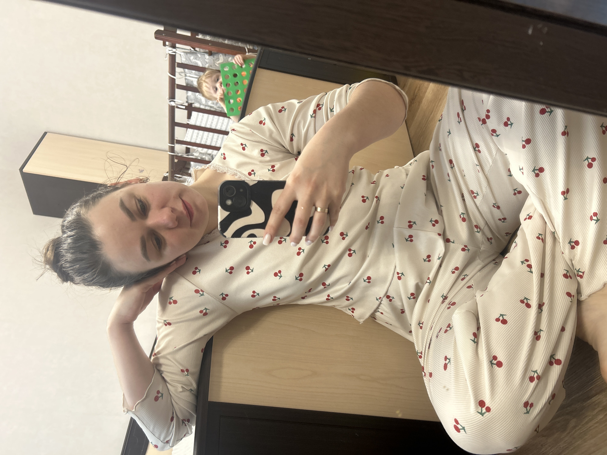 A woman with brown hair in a bun is lying on a wooden surface and taking a selfie with a phone with a black and white swirled case. She is wearing white pajamas with a red cherry pattern. In the background, reflected in a mirror, is a child standing in a crib. The location is Chaikovsky, Russia.