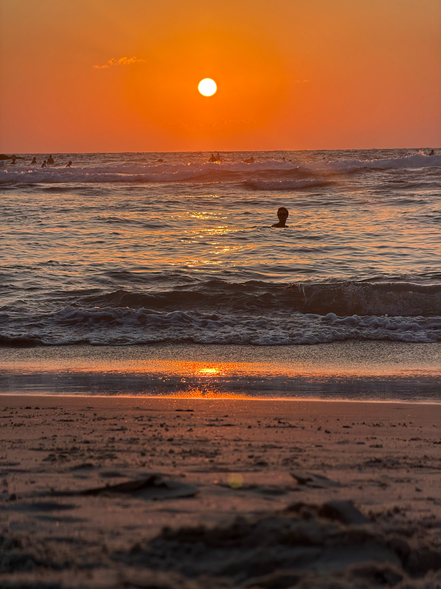 The image depicts a serene beach scene at sunset in Tel Aviv, Israel. The sun, a brilliant orb of orange and yellow, hangs low in the sky, casting a warm, golden glow across the water and sand. Gentle waves lap at the shore, creating a soothing rhythm. In the distance, several people can be seen enjoying the beach, some swimming in the ocean, others wading near the shore. A solitary figure is swimming closer to the foreground, their head just above the water. The sand in the foreground is damp and reflects the sunset's light, creating a shimmering effect. The overall atmosphere is one of tranquility and natural beauty, capturing the essence of a perfect Tel Aviv sunset.