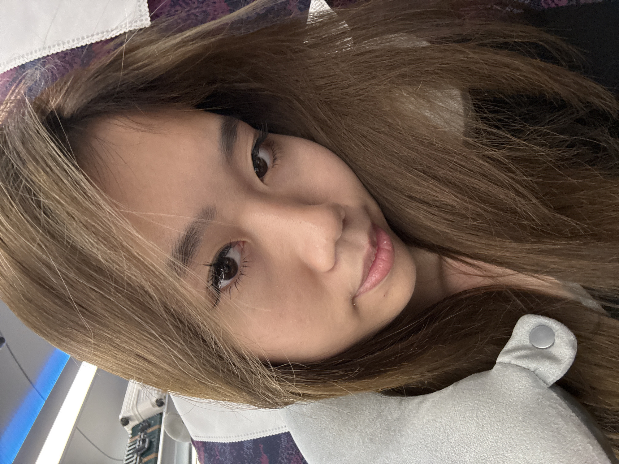 A medium shot of an Asian woman, likely in her early to mid-twenties, with shoulder-length brown hair. She appears to be leaning against a gray pillow. The background features a purple and white patterned surface, potentially a seat. The lighting is diffused and warm. There is no indication of the location being Huashi, China, or any other specific location.