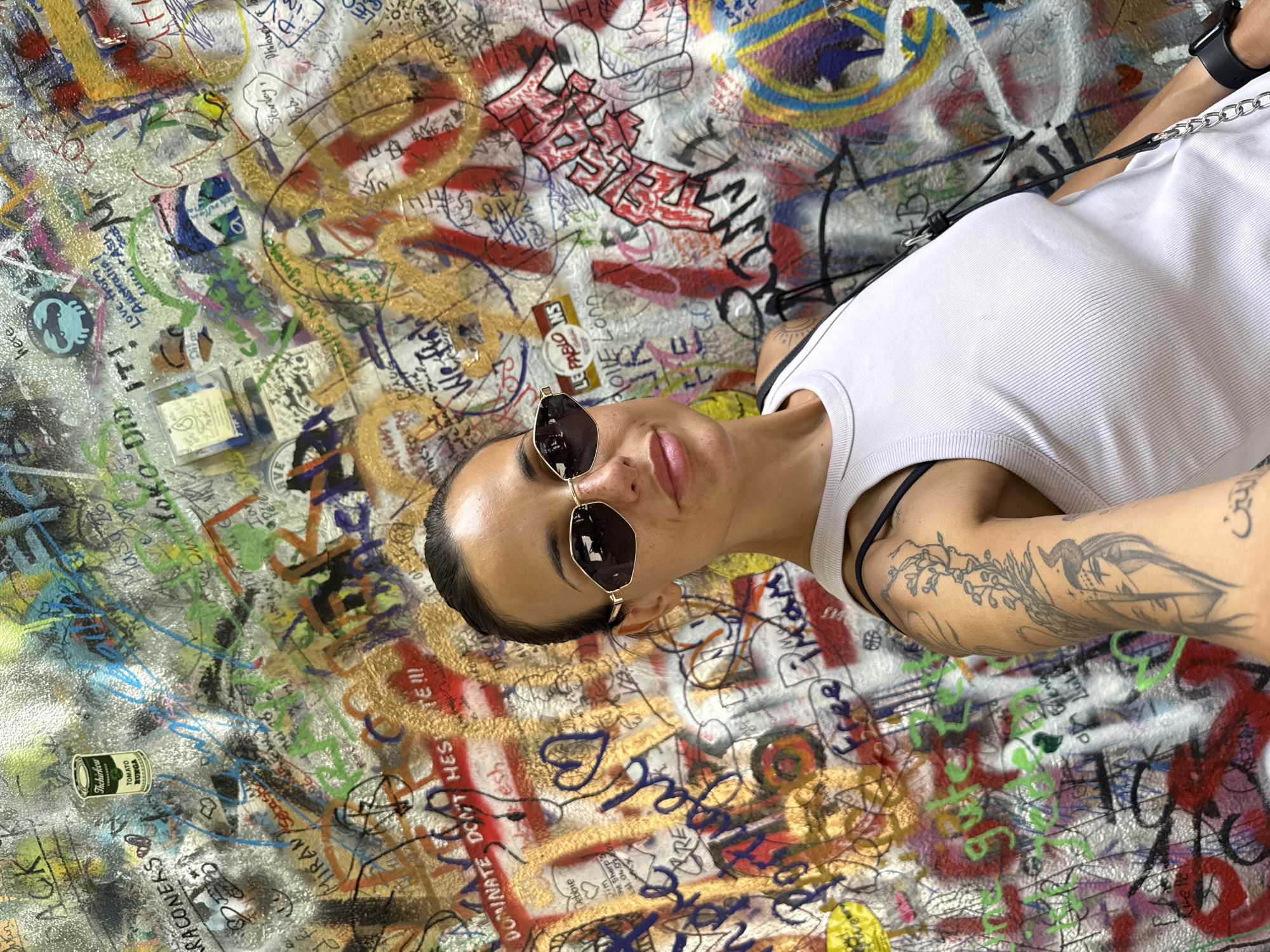 A woman with dark hair and sunglasses is taking a selfie in front of a graffiti-covered wall. She is wearing a white tank top and has tattoos on her left arm. The wall is covered in a colorful array of graffiti, tags, and scribbled messages. This location is identified as the John Lennon Wall in Prague, Czechia. The wall is a popular tourist attraction known for its ever-changing artwork inspired by John Lennon and peace. The image captures the vibrant and dynamic nature of this unique outdoor canvas.