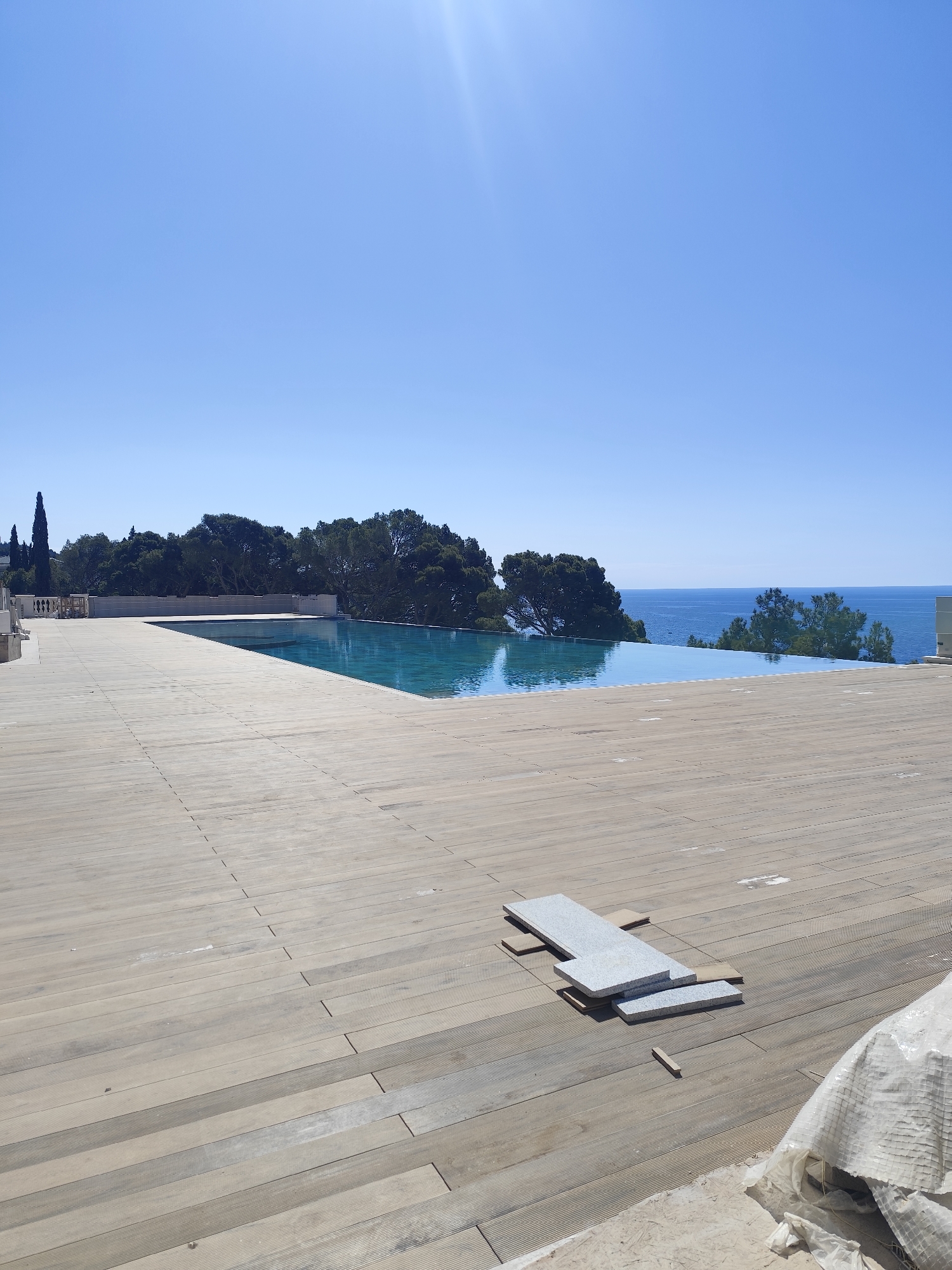 The image shows an infinity pool with a wooden deck surrounding it. The pool seems to overlook the ocean or a large body of water, with trees and other vegetation visible in the background. In the foreground, on the wooden deck, there are several grey stone tiles or pavers stacked on top of each other. To the right of the tiles, there is a large white plastic sheet partially covering something. The sky is clear and blue, suggesting a sunny day. The location appears to be a high-end residential or resort setting. The city and precise location are unknown.