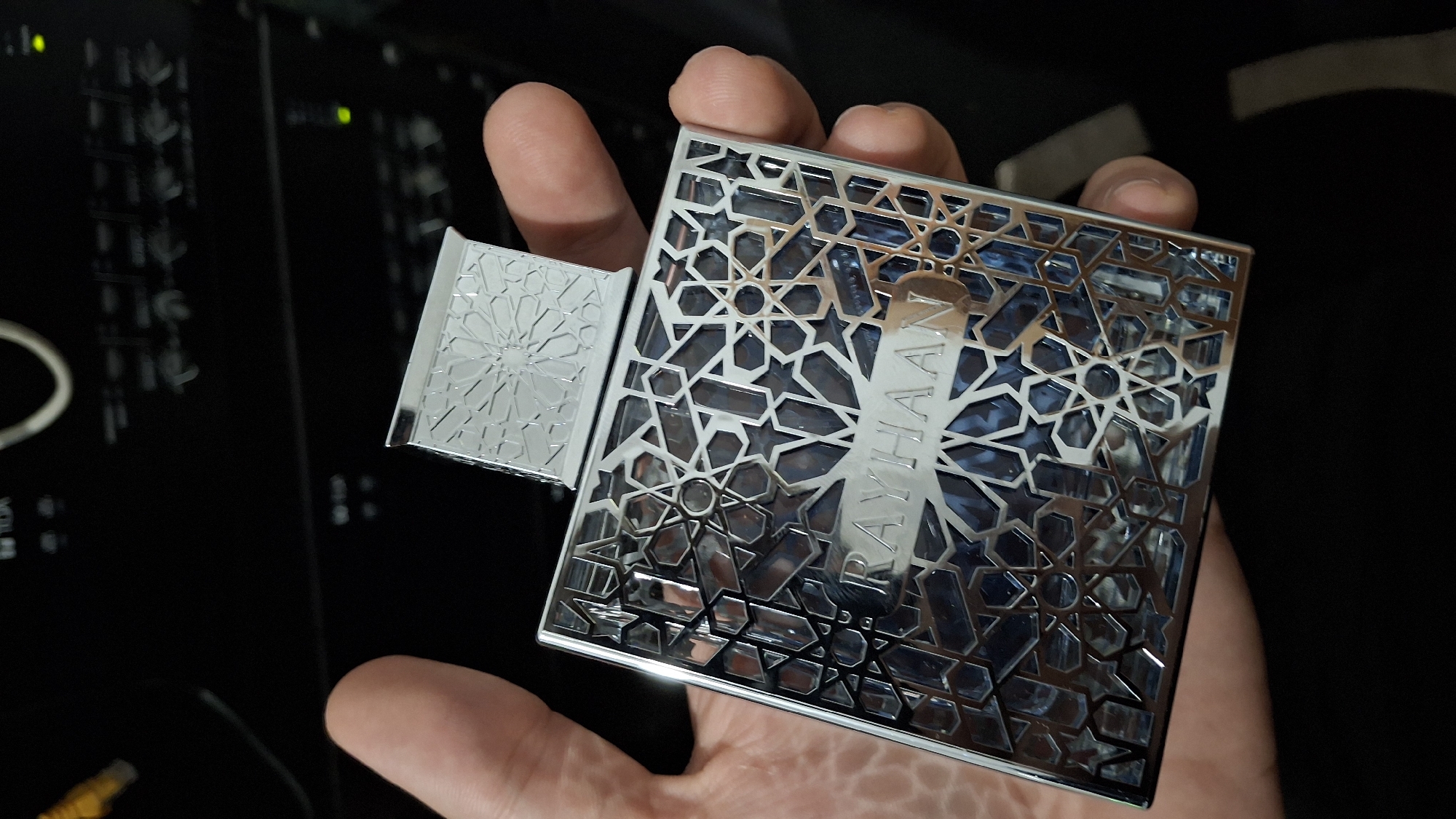A person's left hand holds a silver perfume bottle with an intricate geometric pattern. The bottle has the name "RAYHAAN" etched on it. The background appears to be a black electronic device, possibly audio equipment. The image is a close-up shot, with the focus on the perfume bottle and the hand. The location is not discernible from the image.