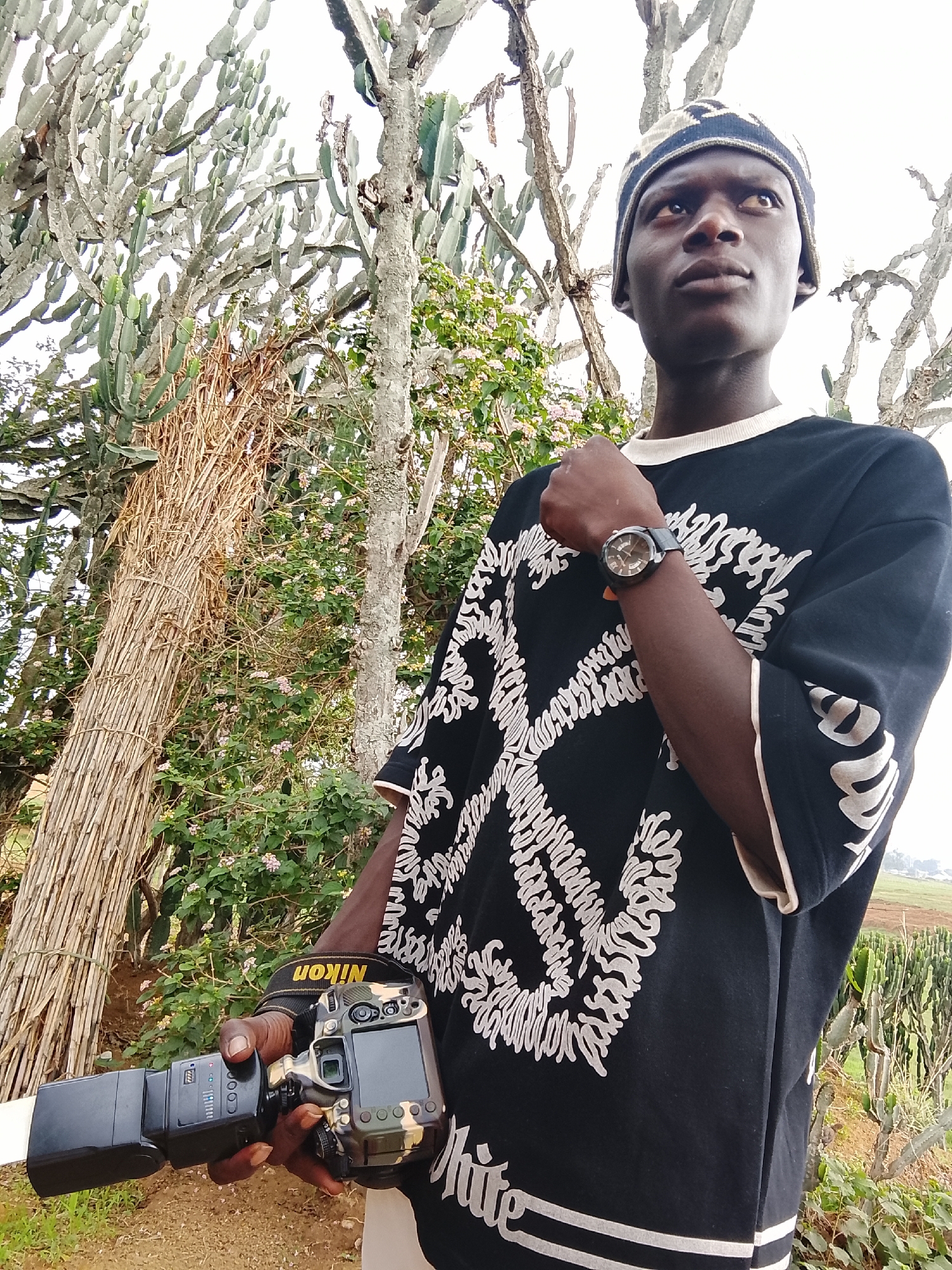 A medium shot depicts a slender African man, dressed in a black and white t-shirt and a patterned beanie, holding a Nikon camera with a flash attached. He's wearing a wristwatch on his left wrist and his left arm is partially raised. The scene is set outdoors amidst a natural environment with tall, slender trees and greenery. In the background, fields or grassy plains stretch into the distance. The location appears to be rural or on the outskirts of a city.