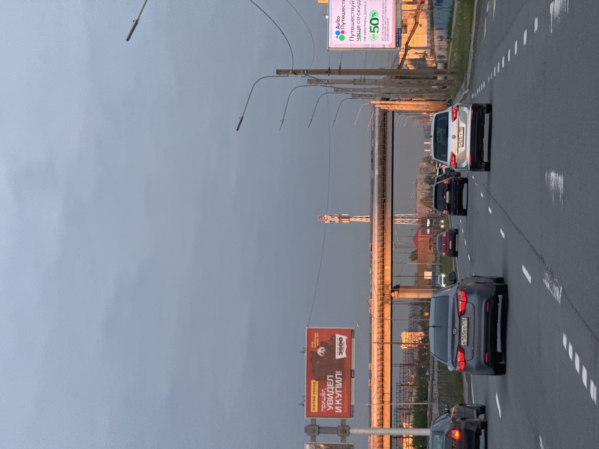 An eye-level shot, rotated ninety degrees clockwise, captures a multi-lane highway or bridge in Saint Petersburg, Russia, during what appears to be late afternoon or early evening. The sky is overcast and hazy, but a warm, golden light from the setting or rising sun illuminates the background bridge structure and distant buildings.
Several vehicles are visible on the road. In the foreground on the right lane, a dark gray Kia sedan with Russian license plate "B145EO 178" is seen from the rear, its brake lights faintly glowing. Further ahead in the same lane is a white Volkswagen van, displaying license plate "A923CT 198". Other vehicles are partially visible in the distance, suggesting moderate traffic. The road features clear white dashed lane markings.
Streetlights line the road, their long, curved arms extending over the asphalt. Two large billboards are prominent features. On the left, a red billboard advertises "КИТАЙ ГОРОД" (Kitay Gorod) with the slogan "пришёл. УВИДЕЛ И КУПИЛ!" (came. Saw and bought!) and features an image of a panda, along with the number "3500". On the right, a multi-colored billboard for "Avito" promotes travel discounts: "Путешествуй чаще со скидкой на квартиры, дома и до 50%" (Travel more often with a discount on apartments, houses and up to 50%).
The background reveals a large bridge structure, possibly a cable-stayed or truss bridge, with numerous supporting elements and visible railings, all bathed in the golden hour light. The license plate regional codes (178 and 198) confirm the location as Saint Petersburg and Leningrad Oblast, Russia.