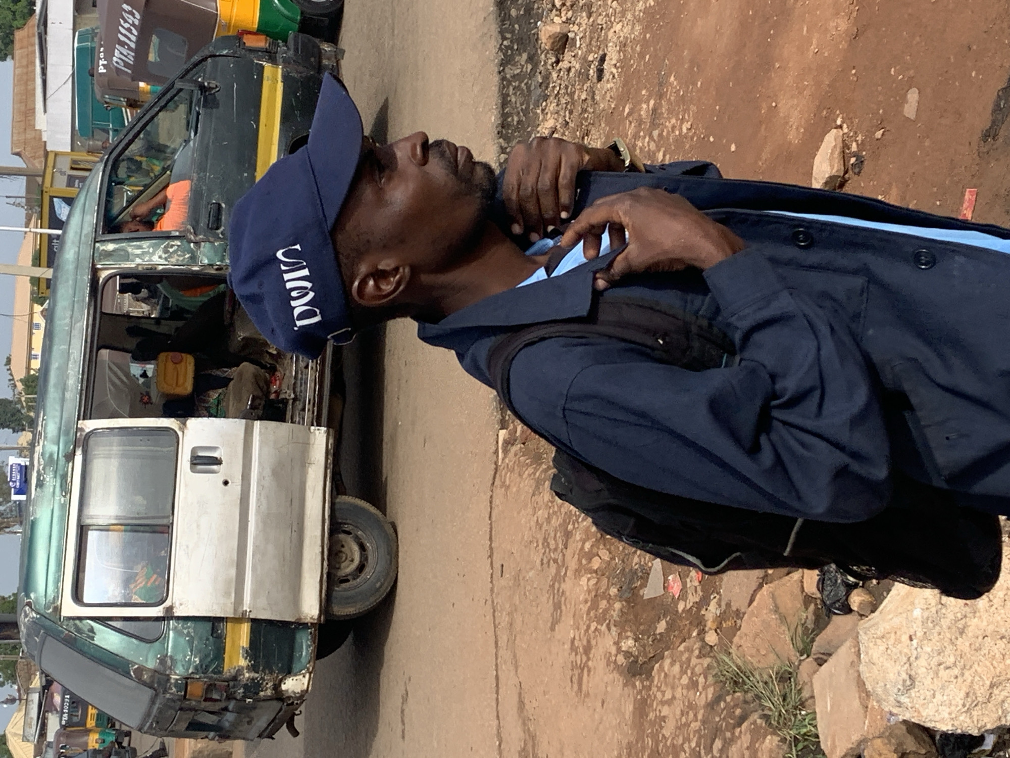 In the city of Gura Top, Nigeria, a man stands in profile, looking to his right. He wears a dark blue uniform jacket over a lighter blue shirt and a matching dark blue baseball cap with white lettering. A black backpack is slung over his shoulders. The man is partially obscuring a weathered green and silver minivan with an open sliding door. Inside the minivan, passengers can be seen, and a man in an orange shirt is visible in the driver's seat. Other vehicles, including an auto-rickshaw with the license plate PTA-11543, are present in the background. The ground is a mixture of reddish-brown dirt and scattered rocks and debris.