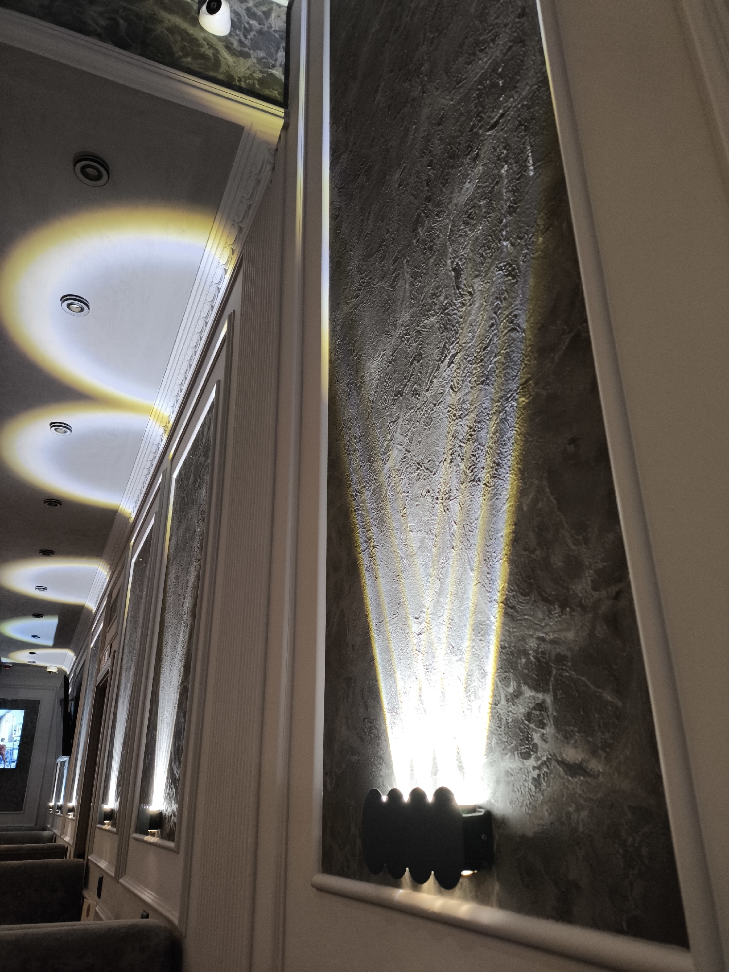 An interior shot captures a well-lit corridor or hallway, likely within a public venue such as a cinema or hotel in Khimki, Russia. The perspective is angled slightly upwards, looking down the length of the passage.
The dominant features are the lighting and architectural elements. The ceiling, painted a light color, is adorned with several recessed spotlights casting distinct, circular glows with a warm, yellowish-white hue. Along the right wall, modern black wall sconces are mounted at regular intervals. These fixtures emit strong, upward-fanning beams of light that highlight the rich, textured surface of the wall, which appears to be dark grey or marble-patterned. The light plays across the uneven surface, creating depth and visual interest.
The left side of the corridor features lighter-colored walls, possibly cream or white, decorated with classical molding and paneling, adding an elegant touch. Further down the hallway on the left, rows of dark, upholstered seating are visible, suggesting a waiting area or lounge. A screen or monitor, displaying an image or video, can be seen in the far background beyond the seating. A white dome-shaped security camera is mounted on the ceiling in the upper left of the frame, near a darker, possibly reflective ceiling section.
There is no specific activity taking place; the scene depicts an unoccupied, illuminated pathway. The artificial lighting suggests an indoor setting, with no indication of the time of day. No visible text is present in the image.