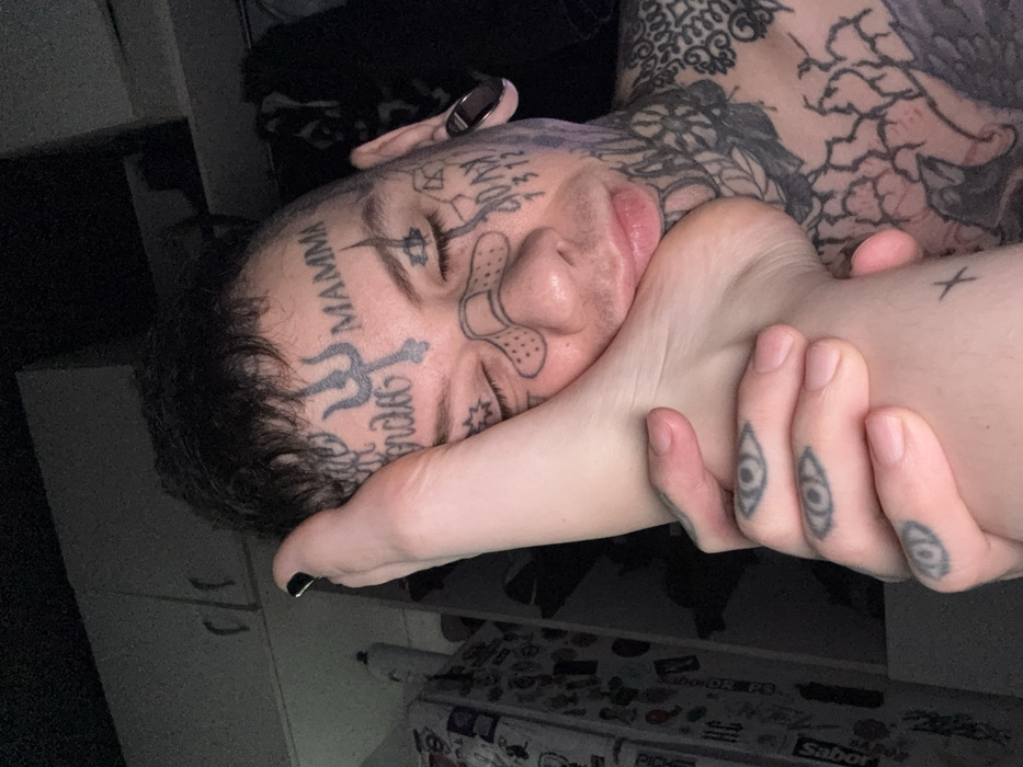 A person with face and body tattoos rests their head on a foot with black nail polish, being held by a hand with eye tattoos on the fingers. The person's face tattoos include the word "MAMMA," a trident symbol, and a drawing of a bandage. The background shows a glimpse of a room with storage and assorted items.