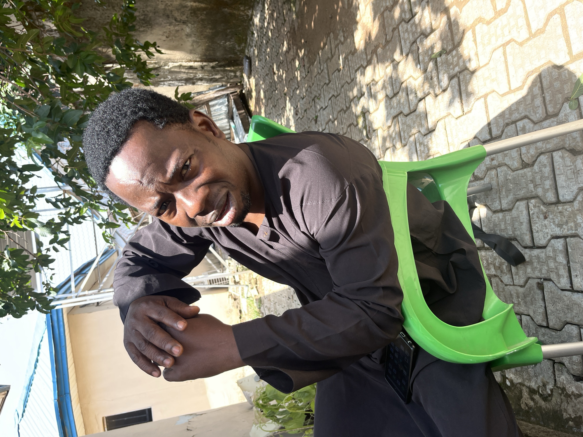 A Black man sits on a bright green plastic chair outdoors in what appears to be a residential area in Abuja, Nigeria. He is wearing a black, long-sleeved robe. His arms are crossed in front of him, with his hands clasped together. He looks towards the camera with a serious expression. A dark-colored smartphone rests on his lap, partially covered by his robe. The ground is paved with interlocking concrete blocks, and patches of sunlight and shadow are visible. Behind the man, a building with a light-colored facade and a blue-trimmed roof is partially visible. Lush green foliage from trees drapes down from above, and parts of a metal structure can be seen. A concrete wall, weathered and showing signs of age, is also present in the background. The scene is brightly lit by natural sunlight.