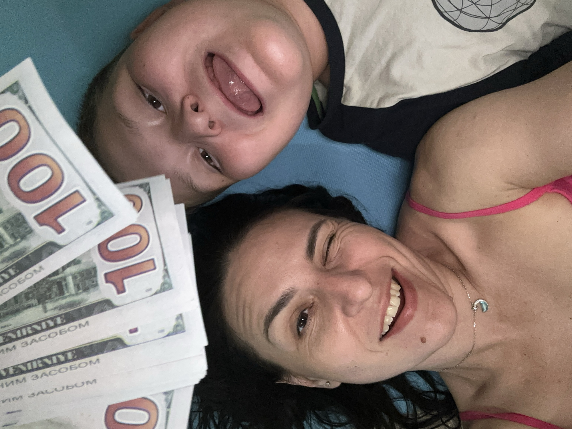 A selfie shows a woman and a child lying down, surrounded by stacks of bills that read "100" in large numbers. The woman, with dark hair and a noticeable mole on her neck, smiles at the camera and winks. She wears a pink top and a delicate necklace with a moon-shaped pendant. To her left, a stack of bills is visible. The child, lying next to the woman, has his mouth open wide, with his tongue visible. The child's shirt has a light-colored top and a darker-colored bottom. The background appears to be a solid blue surface.