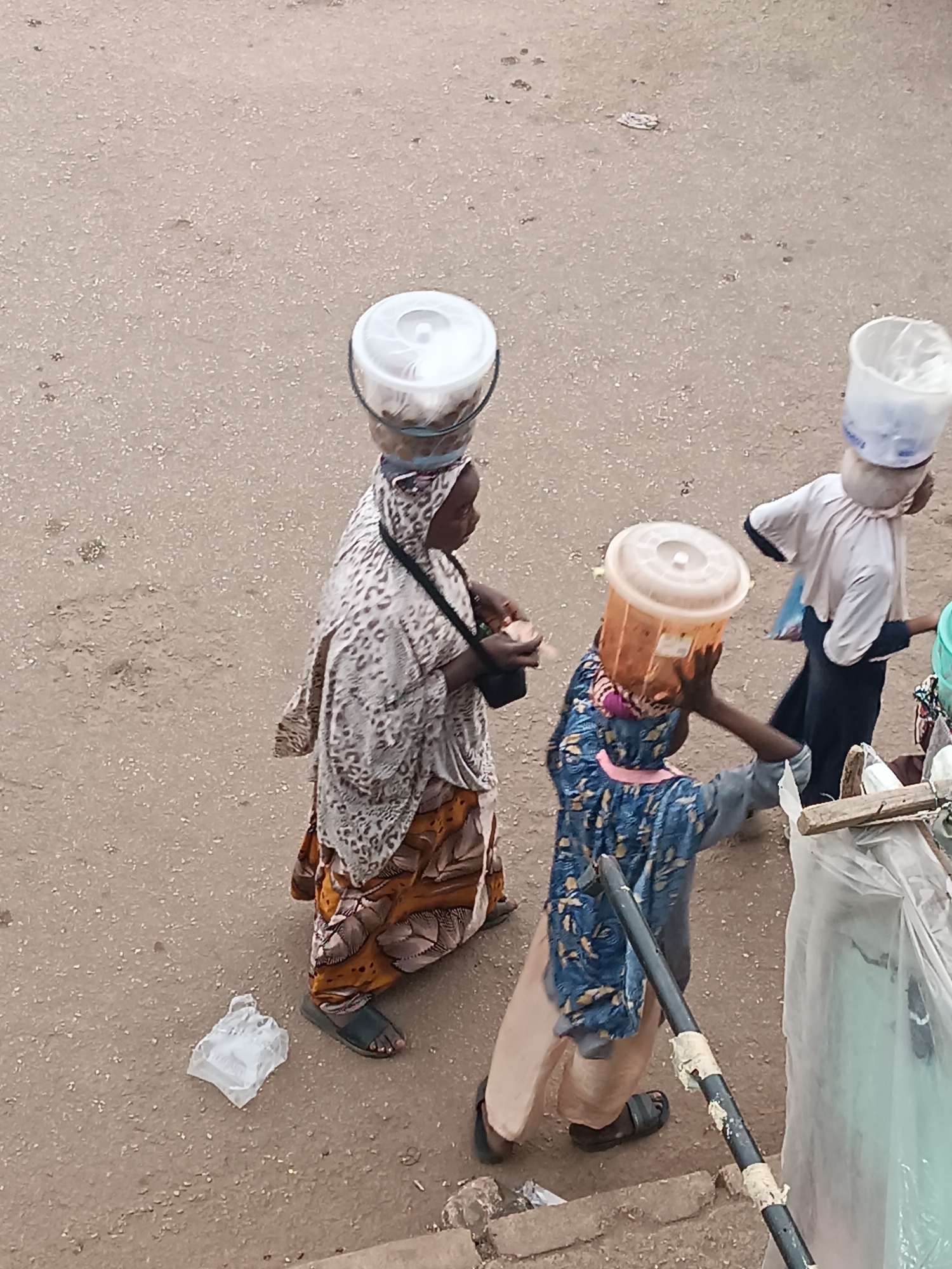 This elevated view captures a street scene in Bukuru, Nigeria, featuring three individuals walking on a light brown, unpaved surface that appears to be dirt or compacted earth mixed with gravel. The subjects are engaged in the common activity of head-carrying goods.
On the left, an adult, likely a woman, is dressed in a patterned white and brown headscarf and a matching long dress. She carries a dark shoulder bag and a clear plastic bucket, filled with unseen items, balanced on her head. Her feet are in dark sandals.
In the center, a young person, possibly a girl, wears a blue patterned top, light trousers, and dark sandals. She uses both hands to steady a translucent orange-brown bucket on her head.
To the right, another young person, likely a boy, is seen in a light-colored top with blue accents and dark trousers. A white plastic bucket, containing what looks like a plastic bag, rests on his head.
The scene is set outdoors during daylight hours. No specific weather conditions are indicated, but the ground appears dry. On the far right, parts of a temporary structure or stall are visible, including a wooden plank, a black metal pole with tape, and transparent plastic sheeting, suggesting a market or roadside commerce. A crumpled white plastic bag lies on the ground near the left-most person. No legible text is visible in the image. The image offers a glimpse into daily life and street activity.