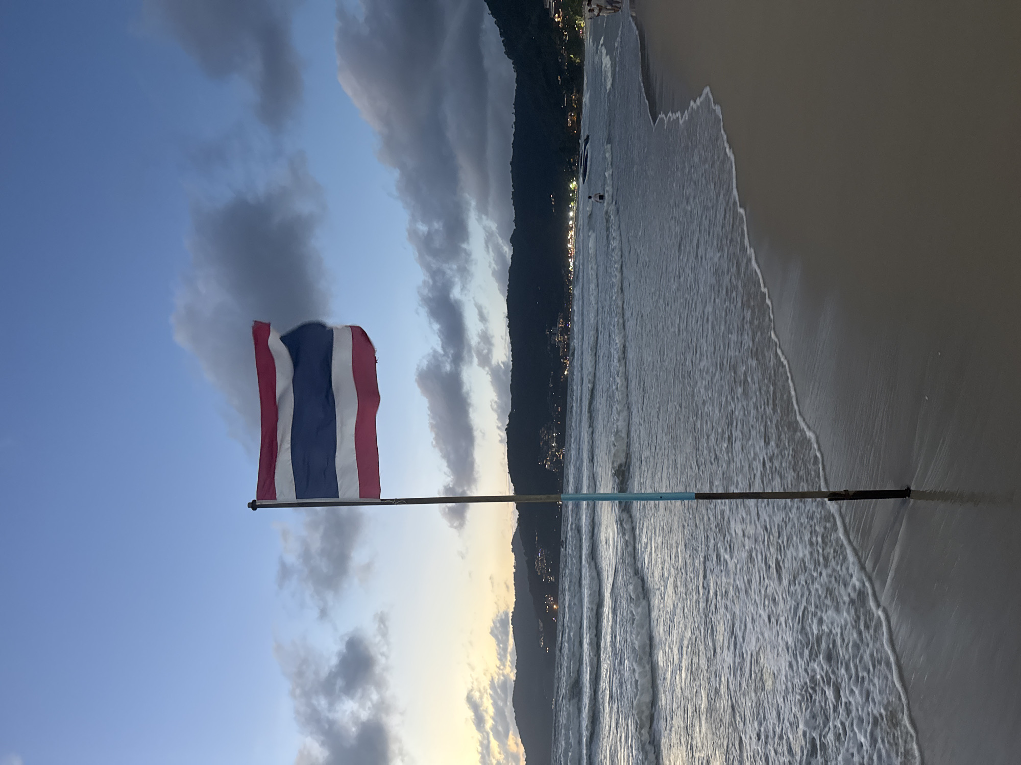 In Patong, Thailand, a Thai flag flies on a pole on a beach during dusk. The flag, with its red, white, and blue stripes, is positioned prominently in the foreground. The ocean is visible with waves rolling onto the sandy shore. In the background, a hazy line of hills dotted with lights suggests a town or city. The sky is a blend of twilight blues and grays, with clouds scattered across it. Several people are present, some walking on the beach and others in the water. The overall scene depicts a serene beach setting at the end of the day.