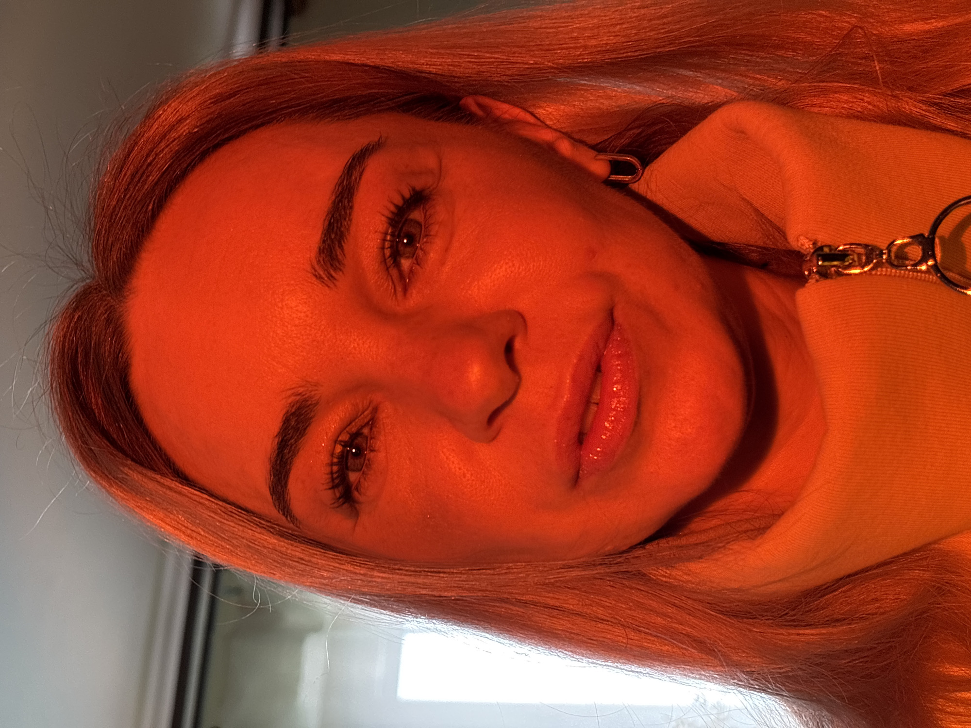 A close-up portrait of a woman with light hair, illuminated by a strong orange light, obscures skin details. The subject is wearing a hooded garment with a visible zipper pull. The background is indistinct. The location and city are unknown.