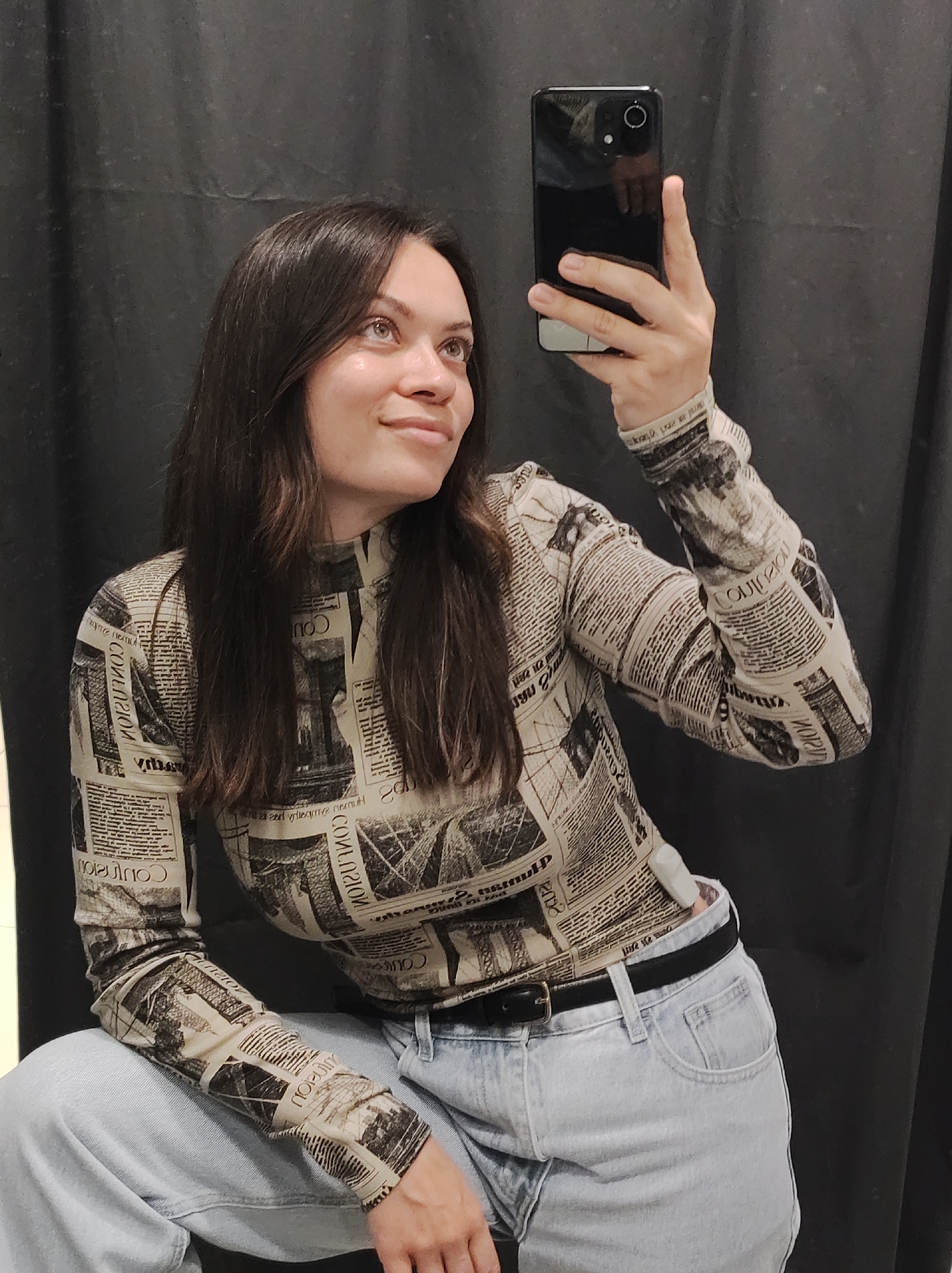 A woman with shoulder-length dark hair is posing for a selfie in front of a black curtain. She is wearing a newspaper-print long-sleeved top and light-wash jeans with a black belt. She holds a black smartphone in her right hand, pointing it at herself. She looks upwards and to the left of the camera. The photograph was taken in a dressing room at a store located at Карла Маркса, 20, Чайковский, Пермский край, Russia, 617763.