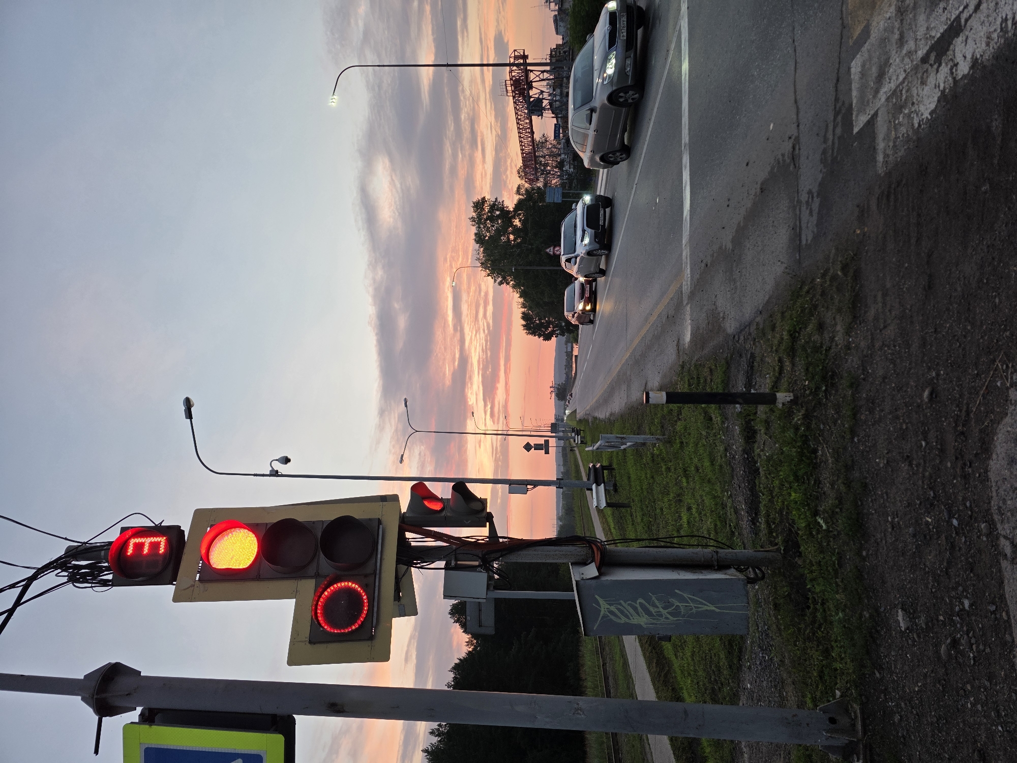 The image depicts a road scene at dusk in Chaikovsky, Russia. A traffic light with red signals, including a countdown timer displaying "3", is prominently featured on the left side of the frame. Several cars are driving on the asphalt road, with a pedestrian crossing marked in white stripes on the right. The sky is painted with the soft hues of sunset, featuring clouds tinged with pink and orange. Trees line the left side of the road, and a power line structure and streetlights are visible in the distance. The overall atmosphere is calm and transitional, capturing the transition from day to night in a semi-urban or industrial area.
