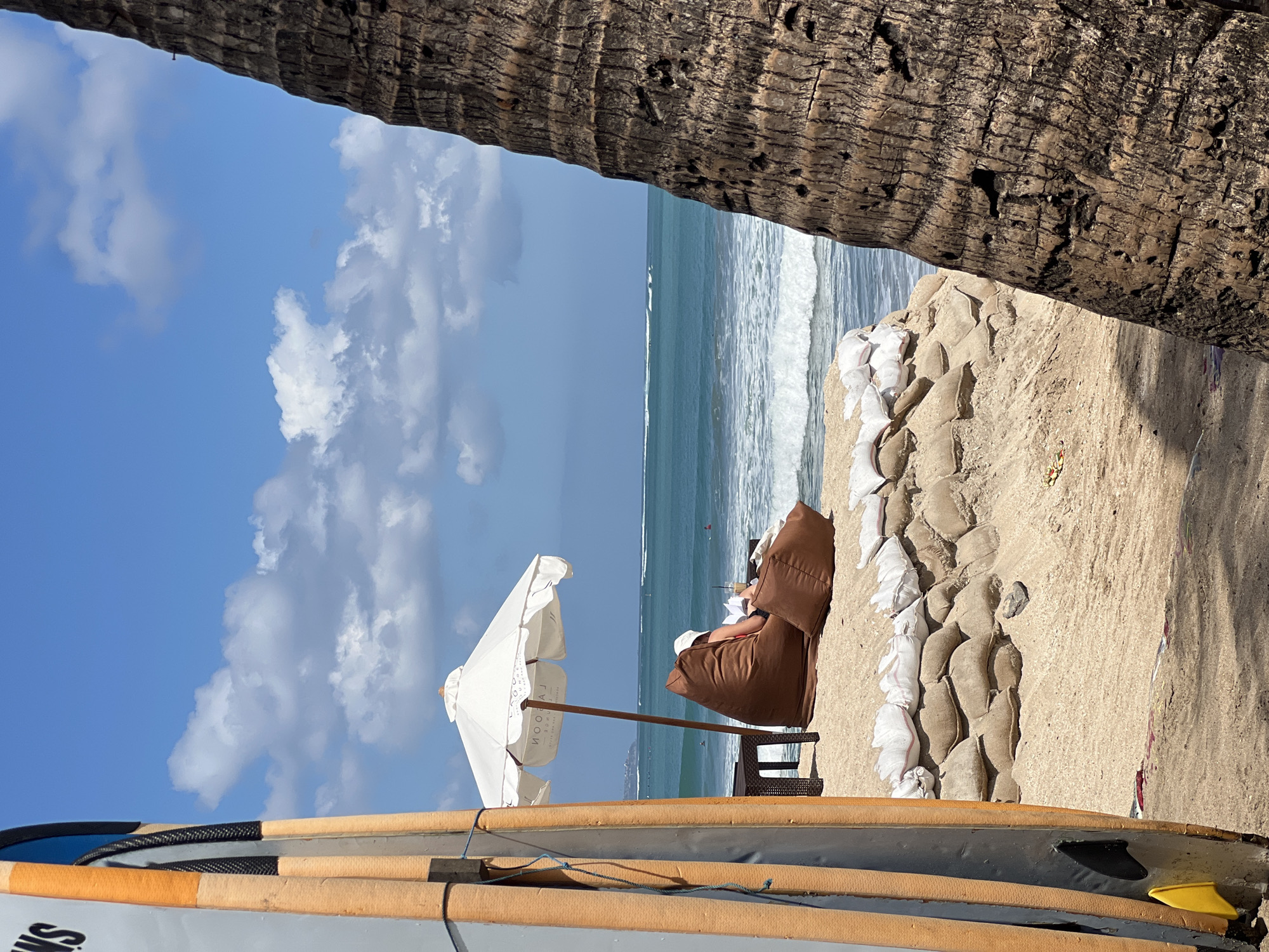 In Kuta, Indonesia, a serene beach scene unfolds under a bright blue sky with scattered clouds. The foreground is dominated by two surfboards leaning against a rough, textured surface, likely a palm tree trunk. Beyond them, a person wearing a white hat and red bikini sits on a large, brown beanbag chair, engrossed in a book and holding a drink. A white umbrella provides shade, and a small table sits nearby. The beach is fortified with sandbags arranged in a tiered pattern, suggesting a coastal defense. The ocean is a vibrant turquoise, with gentle waves rolling towards the shore, and a few distant figures are visible in the water, possibly surfers. The overall atmosphere is one of relaxed leisure and natural beauty.