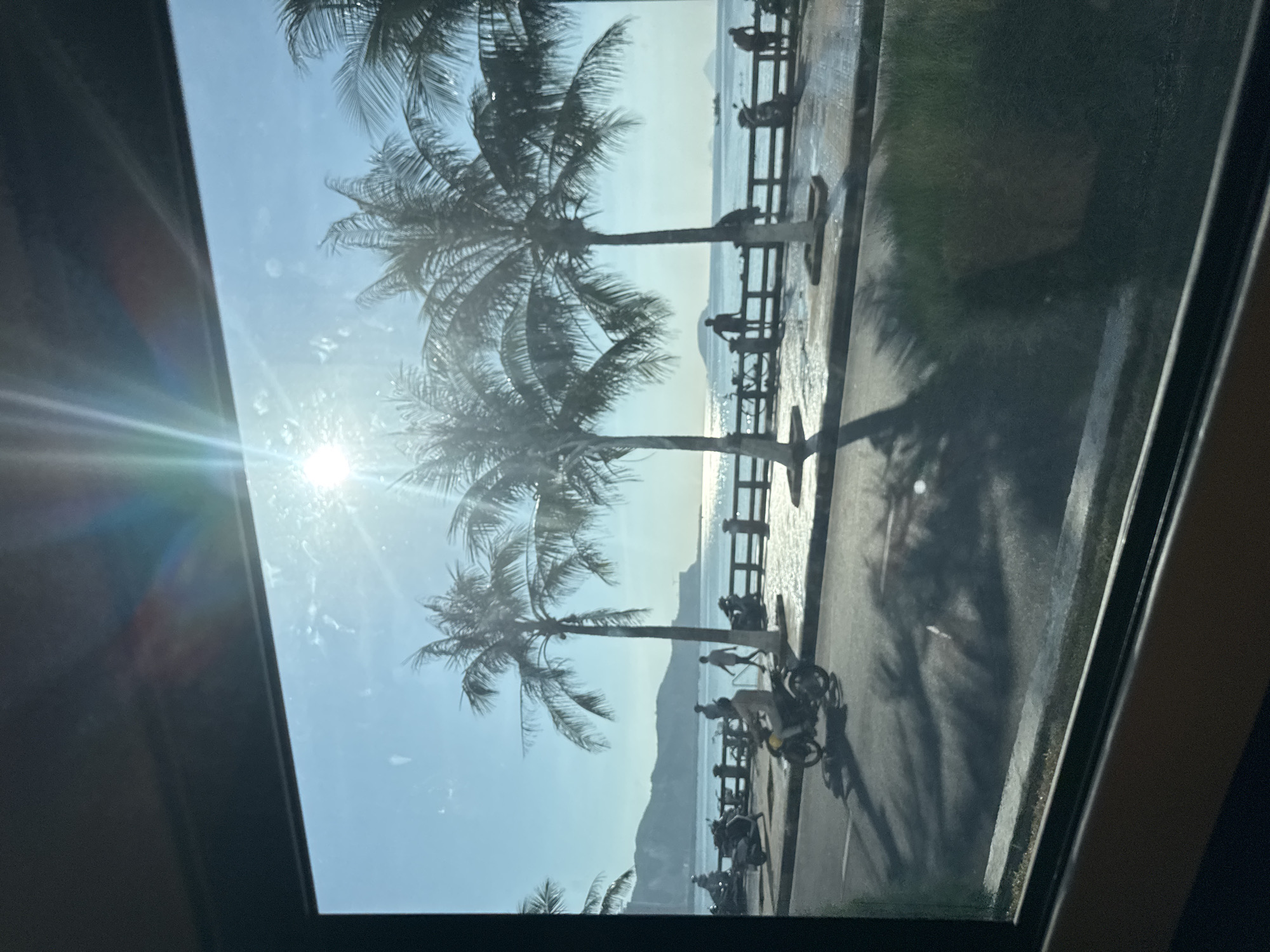 The image depicts a sunny day in Bac Nha Trang Ward, Vietnam, viewed through a window. In the foreground, a street is visible with a person riding a scooter. Palm trees line the street, casting long shadows on the asphalt. Beyond the street is a body of water, likely the ocean, with a promenade and a fence separating it from the street. Several people are seen walking and standing along the promenade. In the distance, mountains and islands can be seen, with the sun shining brightly in the sky, creating a lens flare effect. The overall scene suggests a coastal city with a lively atmosphere.