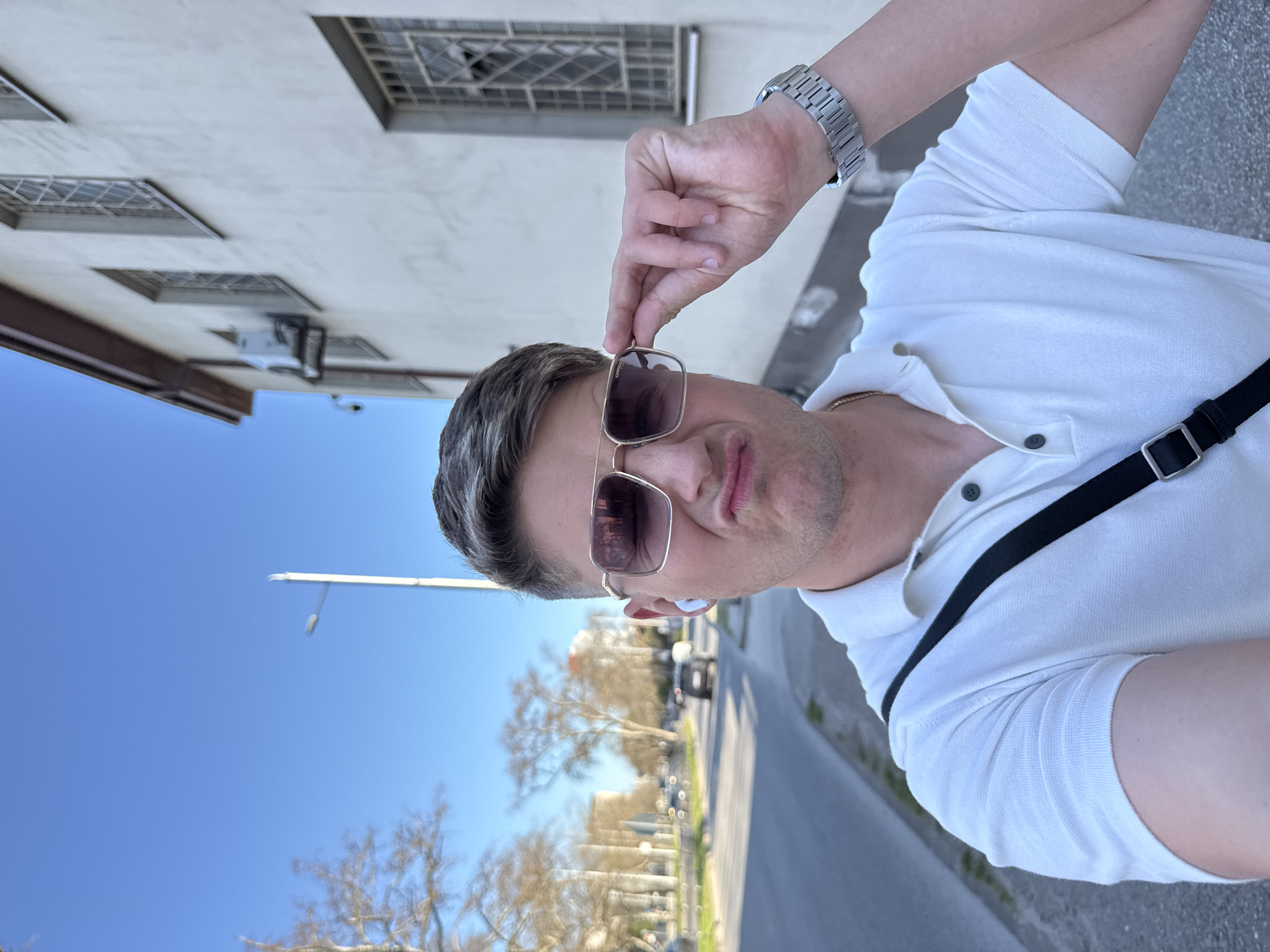 A medium close-up shows a light-skinned man wearing sunglasses with a gold frame and brown lenses. He has short graying hair. The man is wearing a white polo shirt and a silver wristwatch. A black bag strap is visible diagonally across his chest. He is adjusting his sunglasses with his left hand. The background shows a street in Plovdiv, Bulgaria, with trees, a building with barred windows, and a clear blue sky. A street lamp is also visible. The road appears to be curving into the distance. Cars can be seen in the distance.