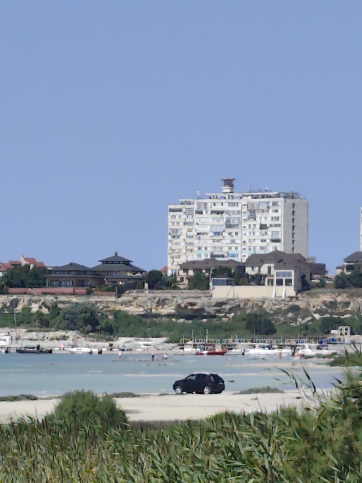 The image depicts a coastal scene in Aktau, Kazakhstan, with a light blue sky dominating the upper portion. In the mid-ground, a multi-story apartment building with numerous windows stands prominently on a hill overlooking the sea. Several other buildings, including what appear to be residential houses with dark, sloped roofs, are situated on the same elevated terrain. A body of water, likely the Caspian Sea, stretches across the lower mid-ground, with several boats docked or anchored near the shore. The water appears calm and light blue, reflecting the sky. The foreground is occupied by a sandy beach with tall, dry grasses swaying in the breeze, creating a natural barrier between the viewer and the sea. A dark-colored SUV is parked on the sand near the water's edge, facing left. The overall impression is of a sunny day at a coastal resort or residential area.
