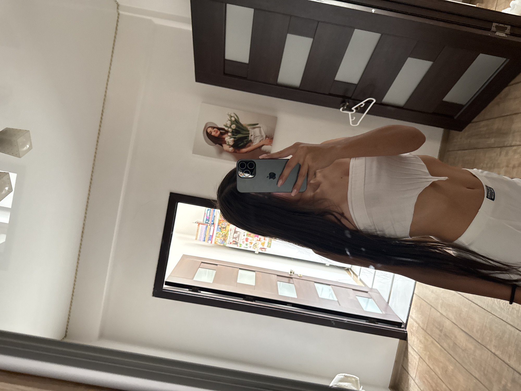 In an indoor setting, a person with long dark hair is taking a mirror selfie. They are wearing a white strapless top and white bottoms. The person is holding a smartphone in their right hand, angled to capture their reflection. Behind them, a dark brown door with frosted glass panels is visible. To the left of the door, there's a white towel hanging on a hook. Further to the left, a doorway leads to another room with shelves filled with books and other items. On the wall to the right of the person, a framed picture of a woman holding a bouquet of white tulips and wearing a hat is displayed. The floor is covered with light brown wooden planks. The lighting suggests an indoor environment, possibly a residential space.