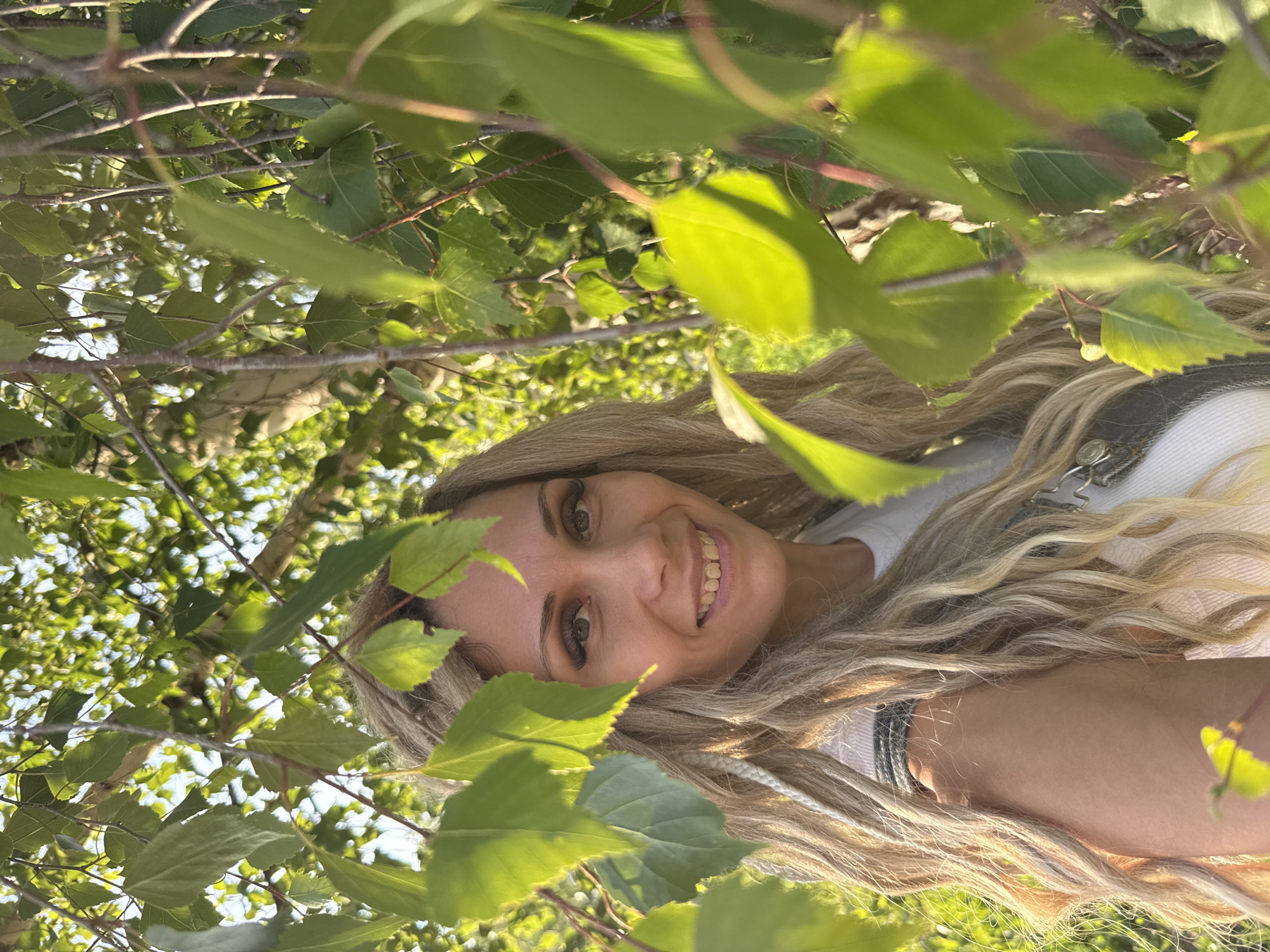 A woman with long, blonde, wavy hair and dreadlocks is partially obscured by green leaves and branches. She is smiling and looking towards the camera. She is wearing a white top and a dark, possibly denim, garment over it. The background consists of blurred green foliage and sunlight filtering through the trees, suggesting an outdoor setting. The image appears to have been taken in Rusanovo, Russia, outdoors.