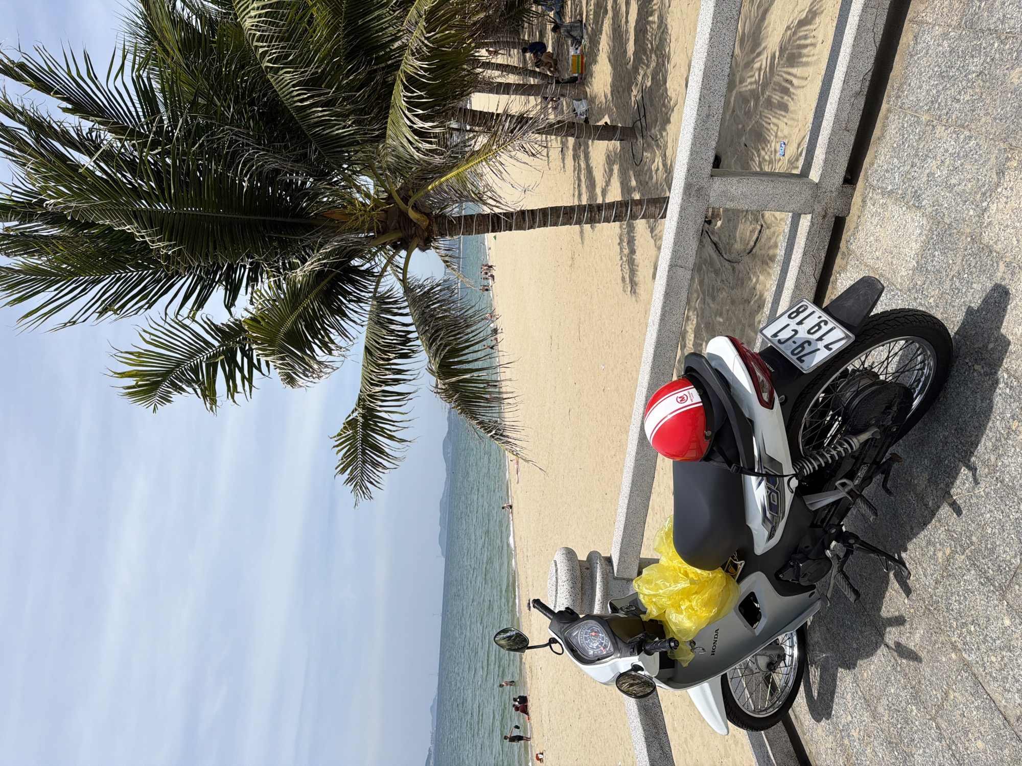A white Honda motorcycle is parked on a paved walkway overlooking a sandy beach in Nha Trang, Vietnam. The motorcycle is parked on the left side of the frame and is partially covered by a bright yellow plastic bag. A red and white helmet rests on the back of the motorcycle seat. Behind the motorcycle, palm trees line the beach, casting shadows on the sand. People are visible on the beach, some swimming in the ocean and others relaxing on the sand. The water is calm, and the sky is partly cloudy. In the background, hills and buildings can be seen in the distance. The scene suggests a typical day at the beach in a coastal city.