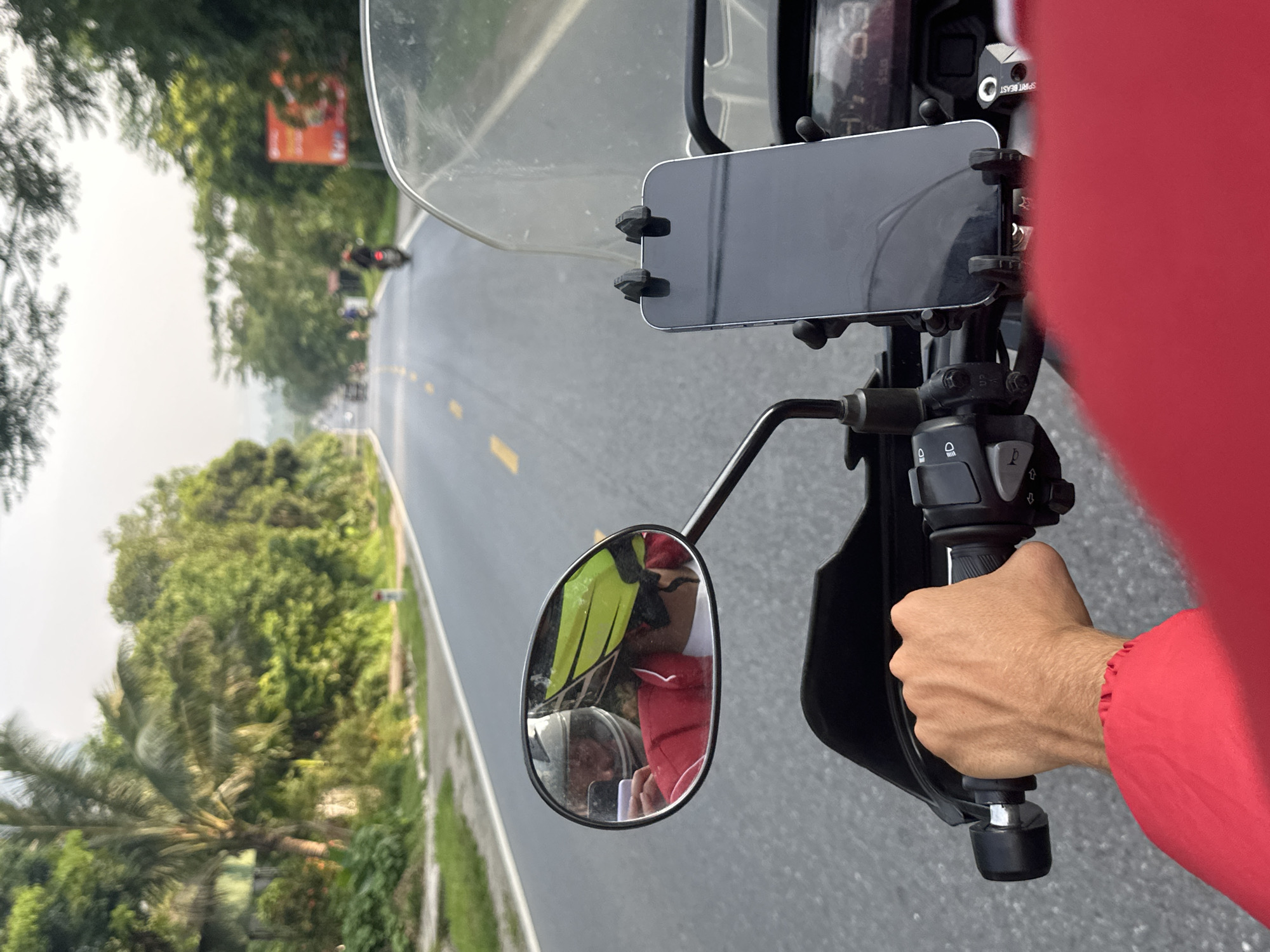 The image provides a first-person perspective from a motorcycle rider traversing a road in Vị Xuyên Commune, Vietnam. The foreground prominently features the right handlebar of the motorcycle, a hand in a red sleeve gripping the throttle, and a smartphone securely mounted vertically above the dashboard. A clear windscreen is also visible.
The road ahead is asphalt, marked with double yellow lines, and appears to stretch into the distance. Several other motorcycles or scooters can be faintly seen in the far background. Lush green trees and dense foliage line both sides of the road, suggesting a tropical or subtropical environment, with a distinct palm tree visible on the left. A blurry orange sign with unreadable text is partially visible through the trees on the right side of the road in the distance.
A rearview mirror, mounted to the handlebar, reflects two individuals. One person is wearing a yellow high-visibility vest, and the other appears to be wearing a red cap or helmet and a red jacket. These are likely passengers or other riders following behind.
Visible text includes the brand "SPIRIT BEAST" on a small metal component near the phone mount, and faint numbers such as "68" and "99" on the motorcycle's dashboard display, possibly indicating speed or mileage. The scene captures an active moment of motorcycling, suggesting travel or a tour on a clear day.