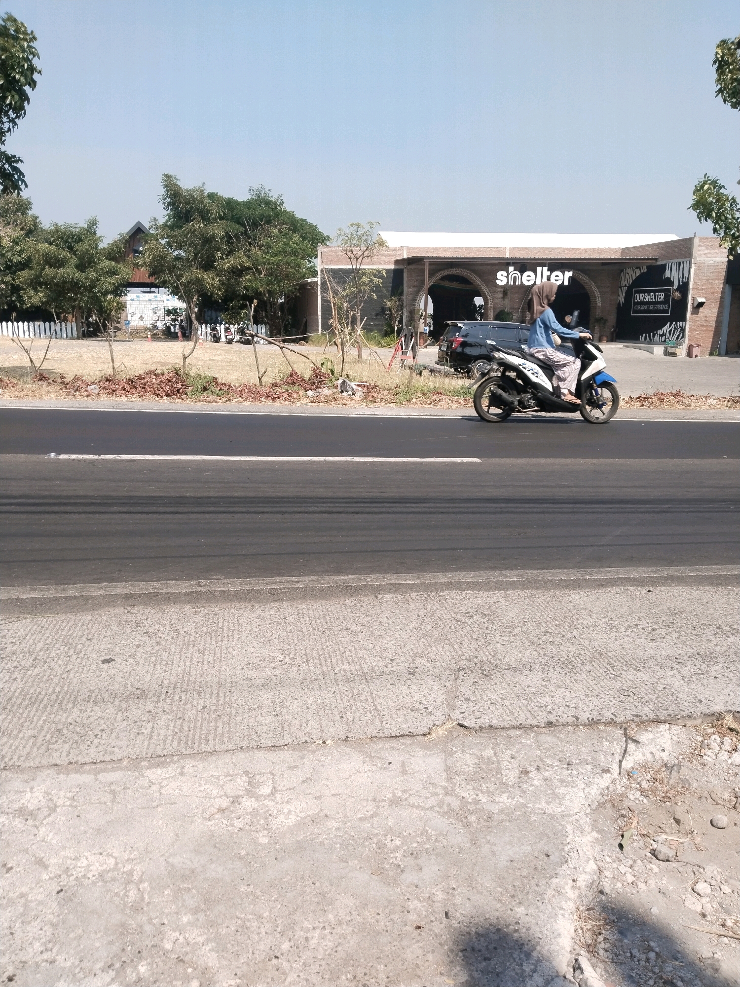 A person is riding a motorcycle on a road in front of a building in Warugunung, Indonesia. The building has "shelter" written on it. The sky is clear and blue. The road is asphalt and the surrounding area is dry and dusty. There are trees and some low vegetation. A black SUV is parked in front of the building.