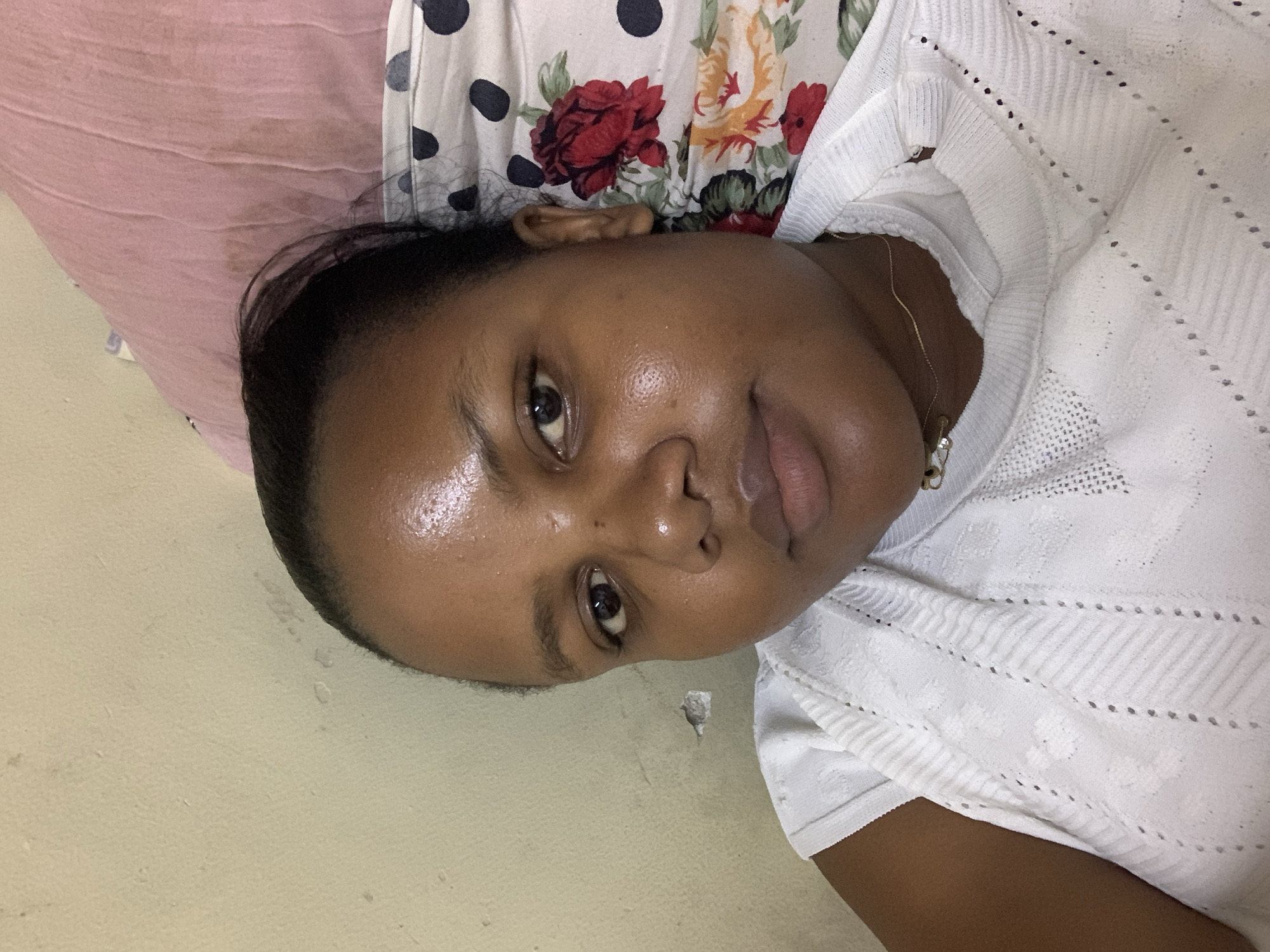 A photograph shows a woman's face and upper body. She has dark hair pulled back from her forehead and is wearing a white top. A gold necklace is visible around her neck. The background includes a pink pillow, and a fabric with floral and polka dot patterns. The setting appears to be indoors, with a beige-colored surface below the woman. There is no information available to determine if this is a photograph that was taken in Dar es-Salaam, Tanzania.