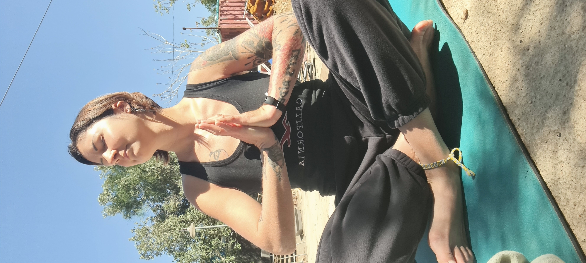 A woman is captured in a serene moment, sitting cross-legged on a turquoise yoga mat outdoors under a bright, clear blue sky. She has short brown hair and her eyes are closed, a calm expression on her face as her hands are pressed together in a prayer-like gesture (Anjali Mudra) at her chest. She wears a black tank top and black sweatpants. Numerous tattoos adorn both her arms, and a black smartwatch is visible on her left wrist. A braided yellow and grey anklet is worn on her right ankle. The black tank top displays the horizontally reversed text "CALIFORNIA" in white, with a red, also reversed, logo beneath it. The background features sparse green trees and shrubs, along with a distant structure, suggesting an outdoor, possibly rural or semi-arid setting like Lachish Regional Council, Israel. The bright sunlight casts distinct shadows on the concrete or dirt ground, indicating daytime. The scene depicts a tranquil yoga or meditation session.