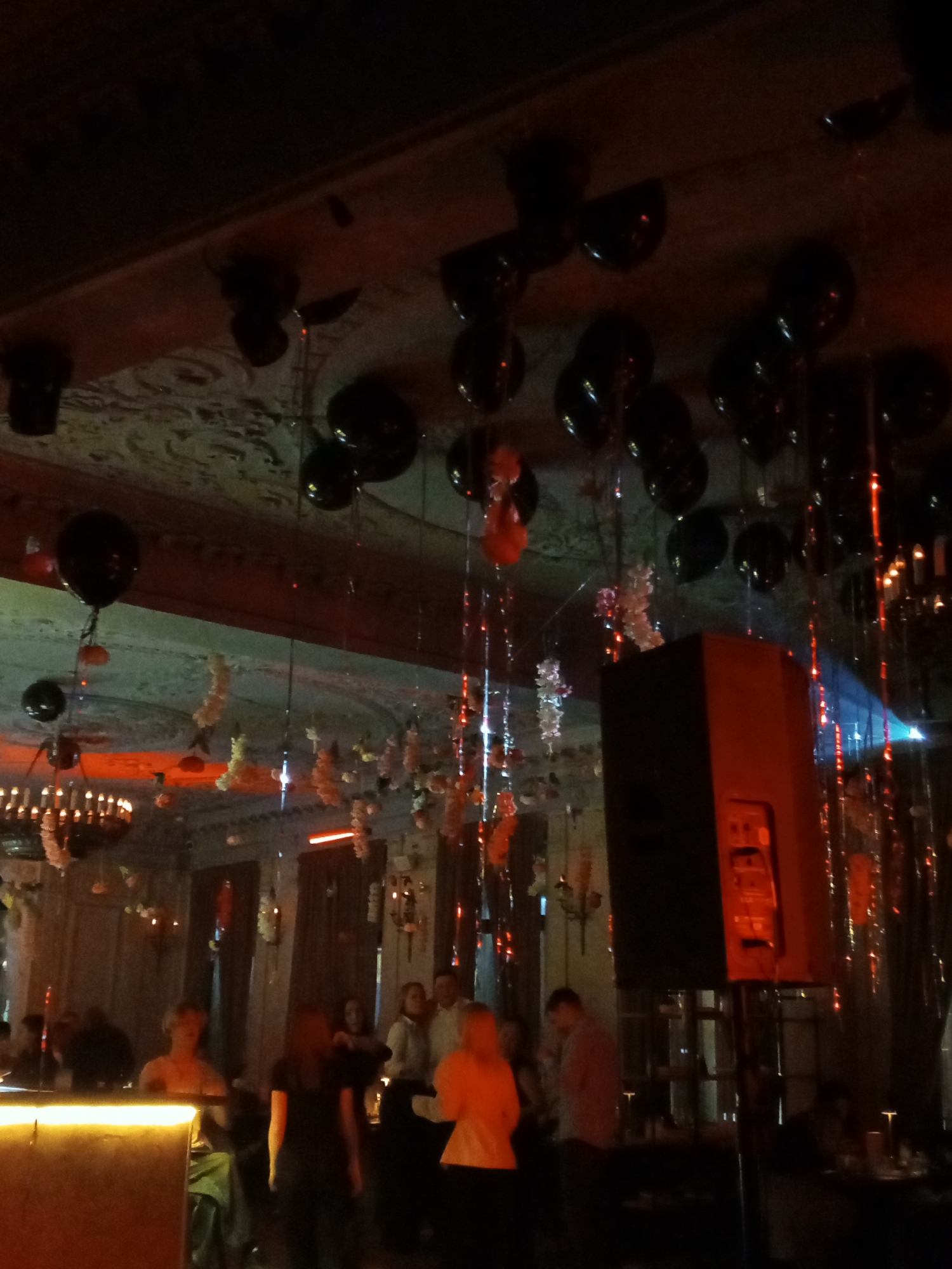 The image captures the ambiance of a dimly lit interior, likely a bar or event space in the city of Saint Petersburg, Russia, decorated for a celebration. A low-lit bar, with patrons conversing, and a sound system with speakers suggest music or a performance will occur. Dark balloons adorn the ceiling alongside dangling decorations and lighting fixtures. The architecture, visible in the ceiling, might suggest a historic building common to the location.