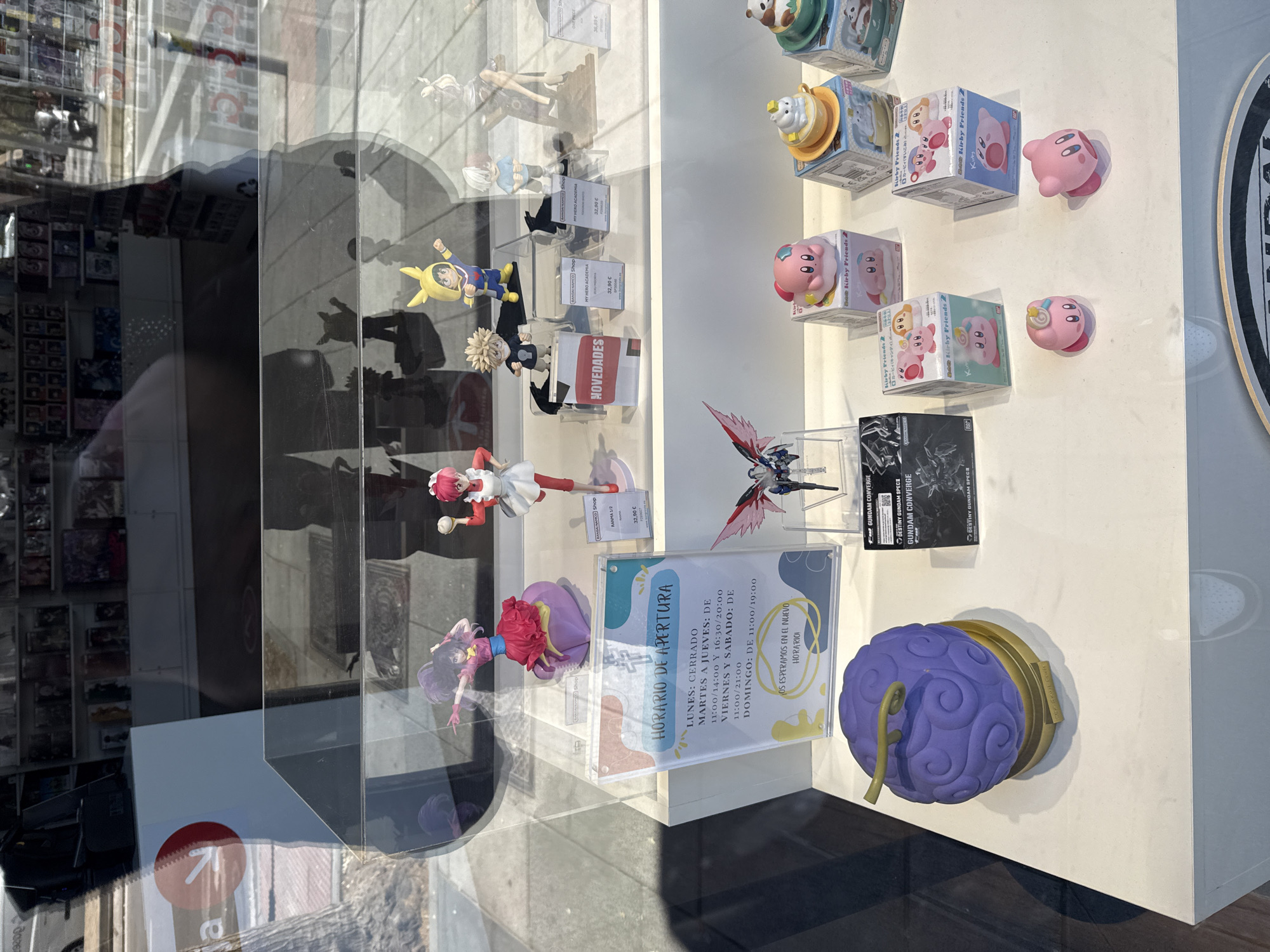 Here is a detailed description of the image:
The image captures a storefront display window, likely in Madrid, Spain. The window showcases a collection of anime and manga figurines, toys, and collectibles. Several shelves are visible, displaying the items.
Featured items include figurines from "My Hero Academia" and "Ranma 1/2", along with "Kirby" toys in boxes, and Gundam kits. A sign displays the store's opening hours (HORARIO DE APERTURA) written in Spanish.
The outside of the window reflects the streetscape, suggesting an urban location. The window's reflection shows parts of buildings and the sidewalk.