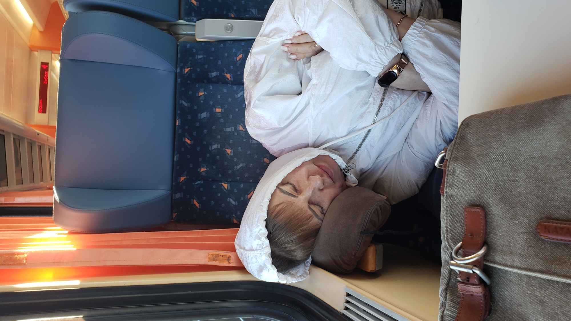 The image shows a woman asleep on a train, leaning her head on a brown pillow. She is wearing a white hooded jacket and her arms are crossed. A watch is visible on her left wrist. In the foreground, a brown canvas bag with leather straps is resting on a table. The train seats in the background are blue with a patterned fabric. The window shows orange curtains and sunlight streaming in, indicating it is daytime. Above the seats, a digital display shows "VÝCHOD: Košice 4". This train is likely traveling from Spišská Nová Ves, Slovakia, as indicated by the destination "Košice" on the display and the general context of the location.