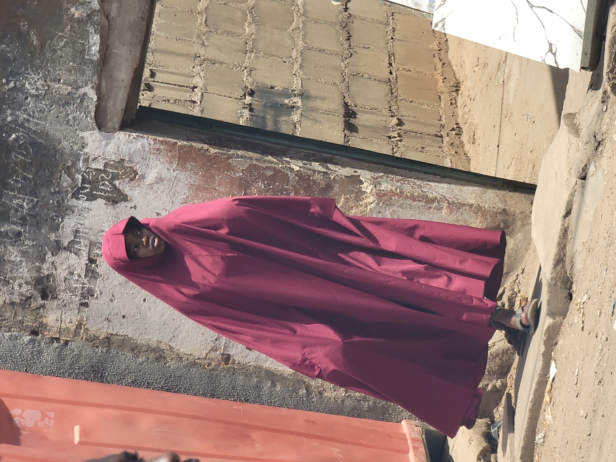 This outdoor image, captured in Maiburiji, Nigeria, features a person standing against a backdrop of weathered walls and ground. The subject, likely a young person, is positioned centrally and looks directly at the viewer with a neutral expression. They are wearing a flowing, full-length garment, probably a hijab or abaya, in a deep maroon color, which covers their body entirely except for their face. Visible on their right foot is an open-toed sandal with light brown and possibly blue-grey straps.
The scene depicts a sunny day, indicated by the bright, harsh lighting and distinct shadows cast on the surfaces. The weather appears clear. The background consists of various architectural elements and terrain. To the immediate left of the person, there is a rough, textured wall, possibly concrete or plaster, showing signs of age with peeling white paint over a darker, greyish-black base. Faint, irregular markings or script, possibly Arabic or informal graffiti, are visible on this wall. Above and behind the person, a section of a light brown brick wall with dark mortar lines is visible, suggesting a building structure. To the right, the ground is sandy and uneven, leading to a raised concrete or stone edge. A partial view of a reddish-orange container or object is seen in the lower left corner. The overall setting suggests a rustic or unpaved area within a town or village.